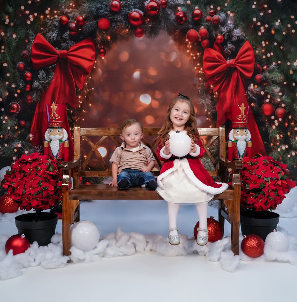 Children seated on wooden bench against festive Christmas backdrop with red ornaments and bows.