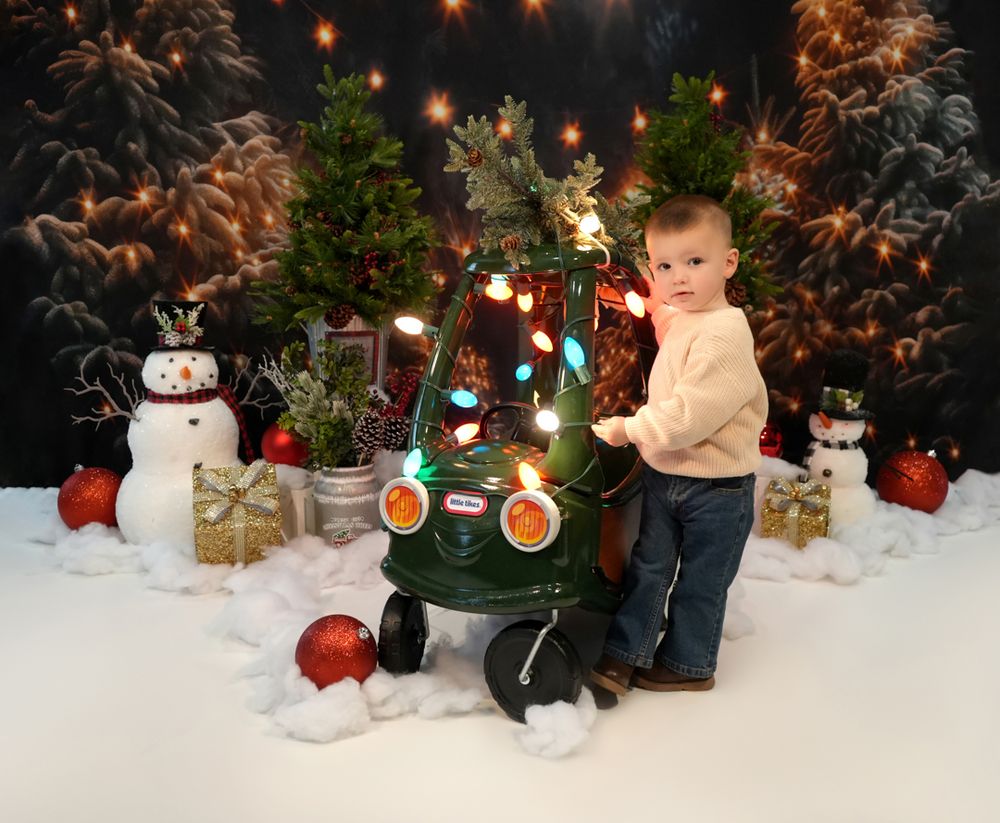 A festive winter photo session setup with Christmas trees, snowmen, and twinkling lights in a cozy indoor studio setting.