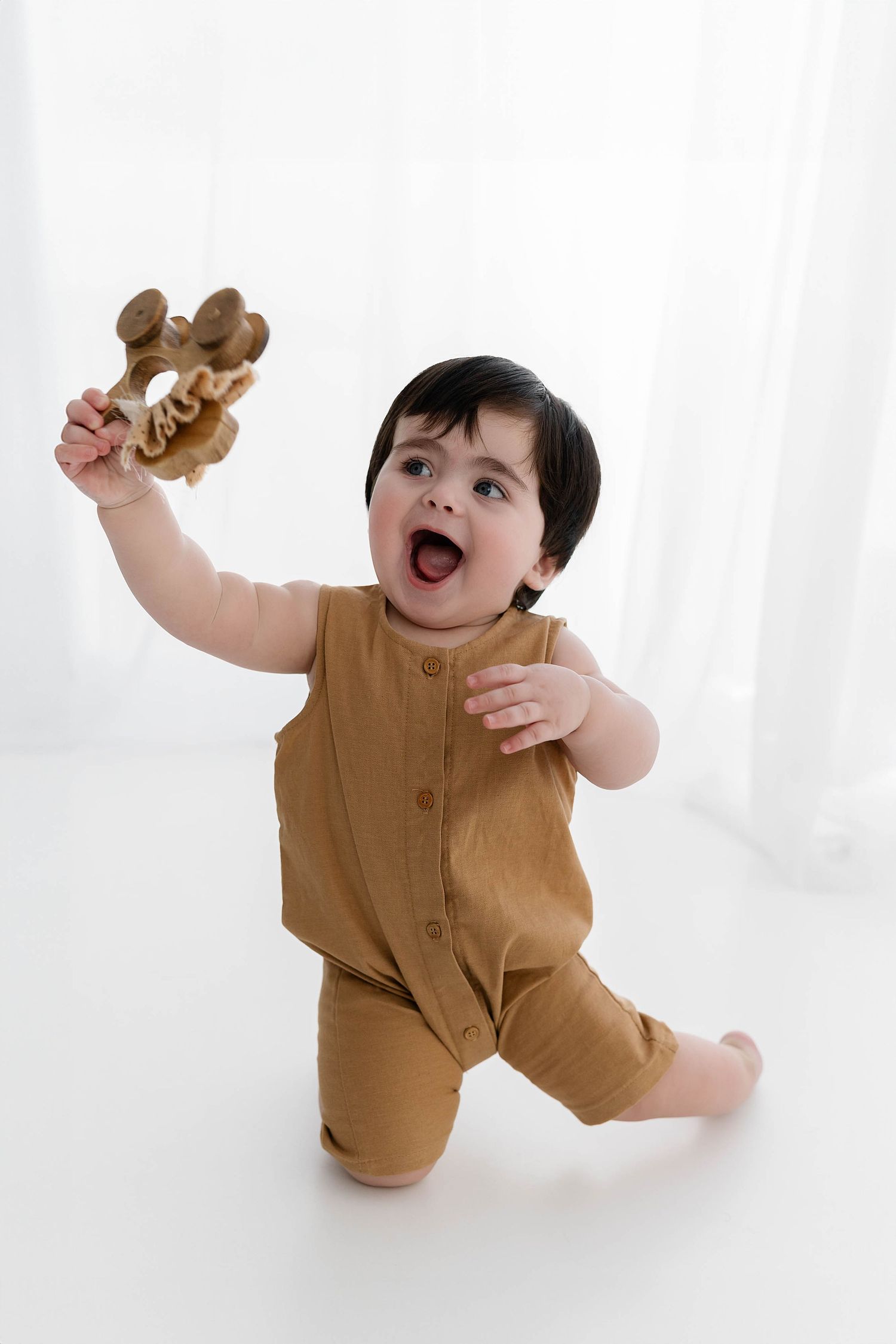 Excited toddler in brown romper holding up wooden toy while sitting against white background.