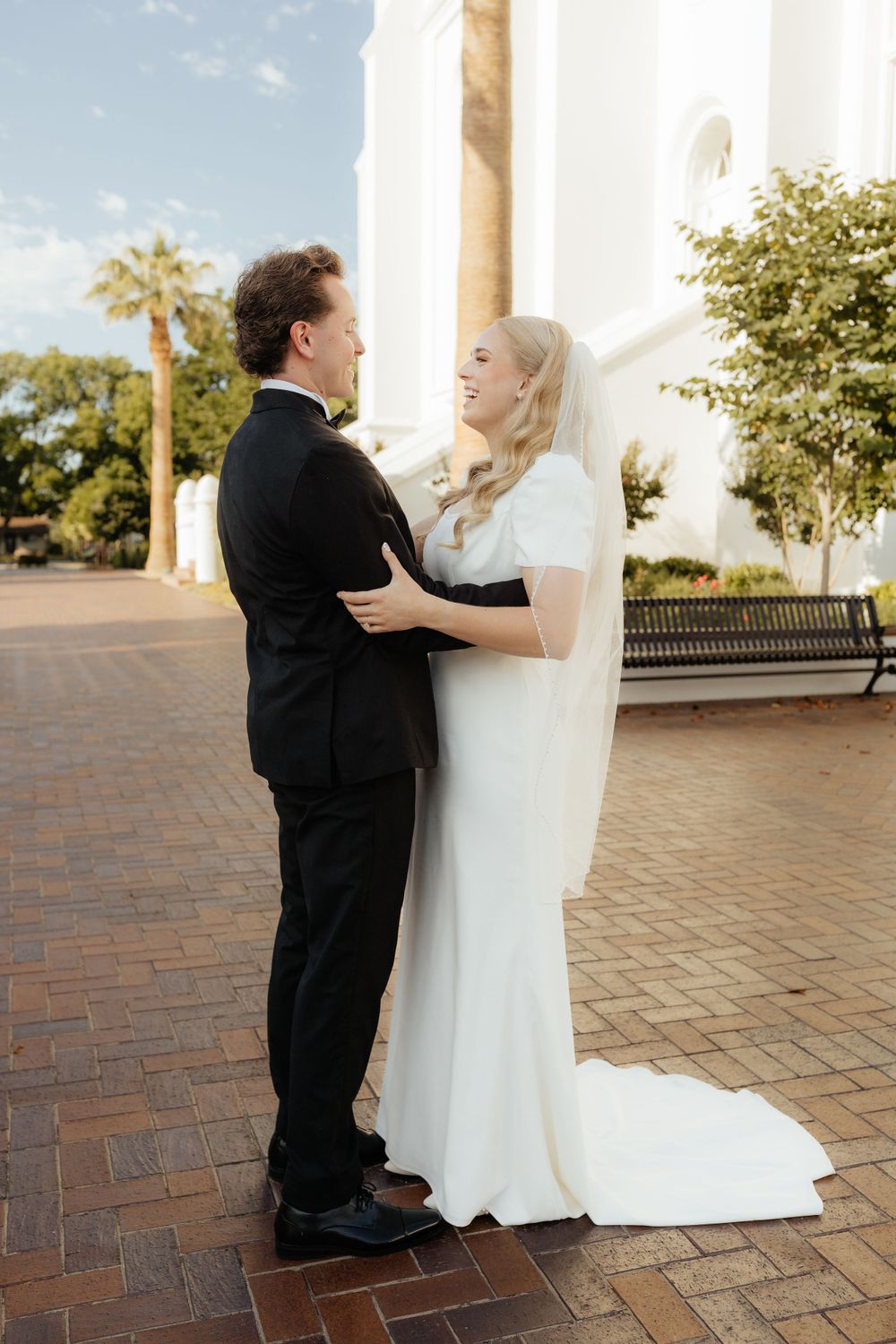 Bride in white gown and veil smiles at groom in black tuxedo while embracing near white building.