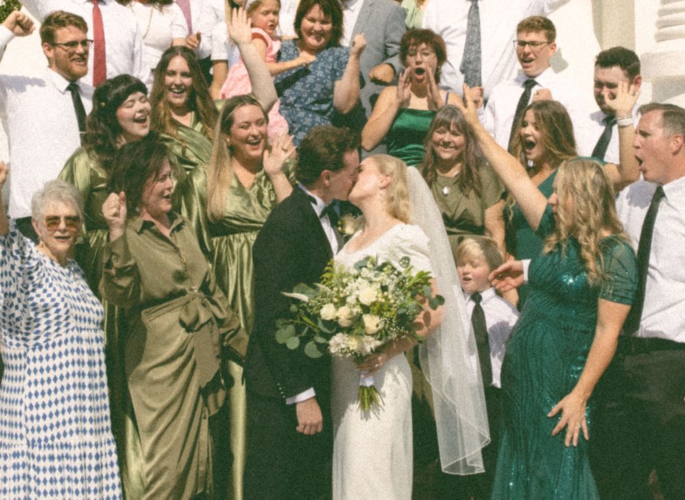 Joyful wedding party celebrates as bride and groom share a kiss on church steps.
