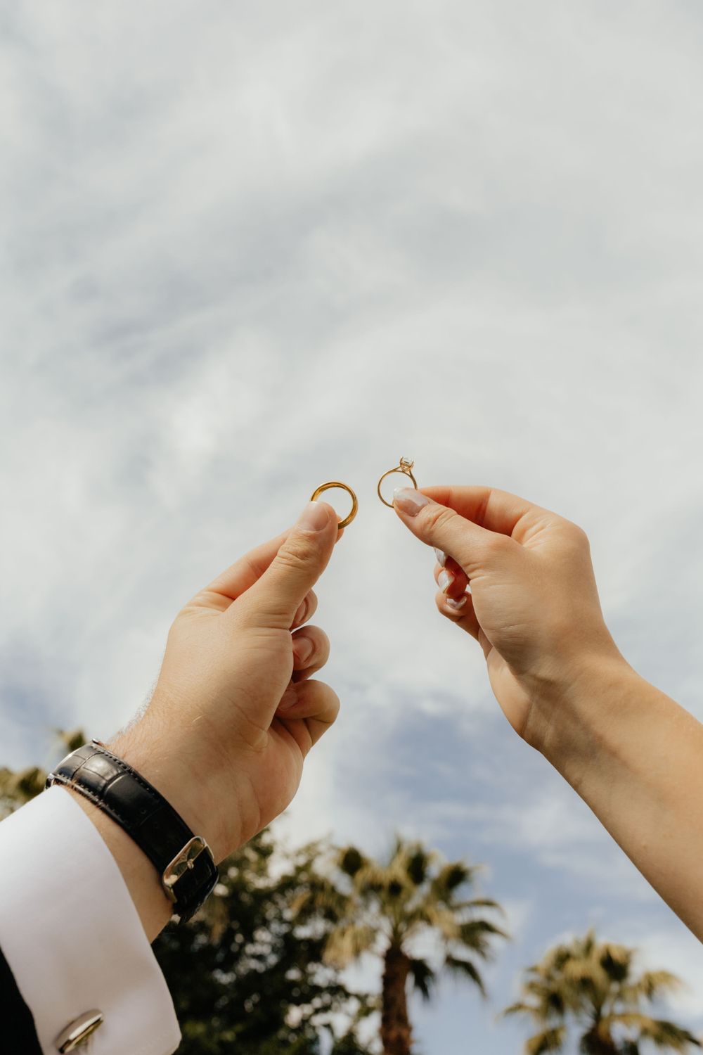 Bride and groom holding up matching gold wedding rings against a cloudy sky.
