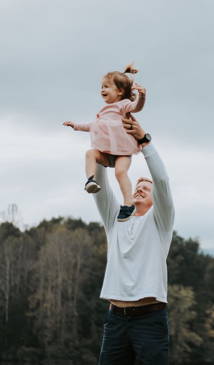 A joyful moment of play as a child in a pink dress is lifted high against a cloudy sky.