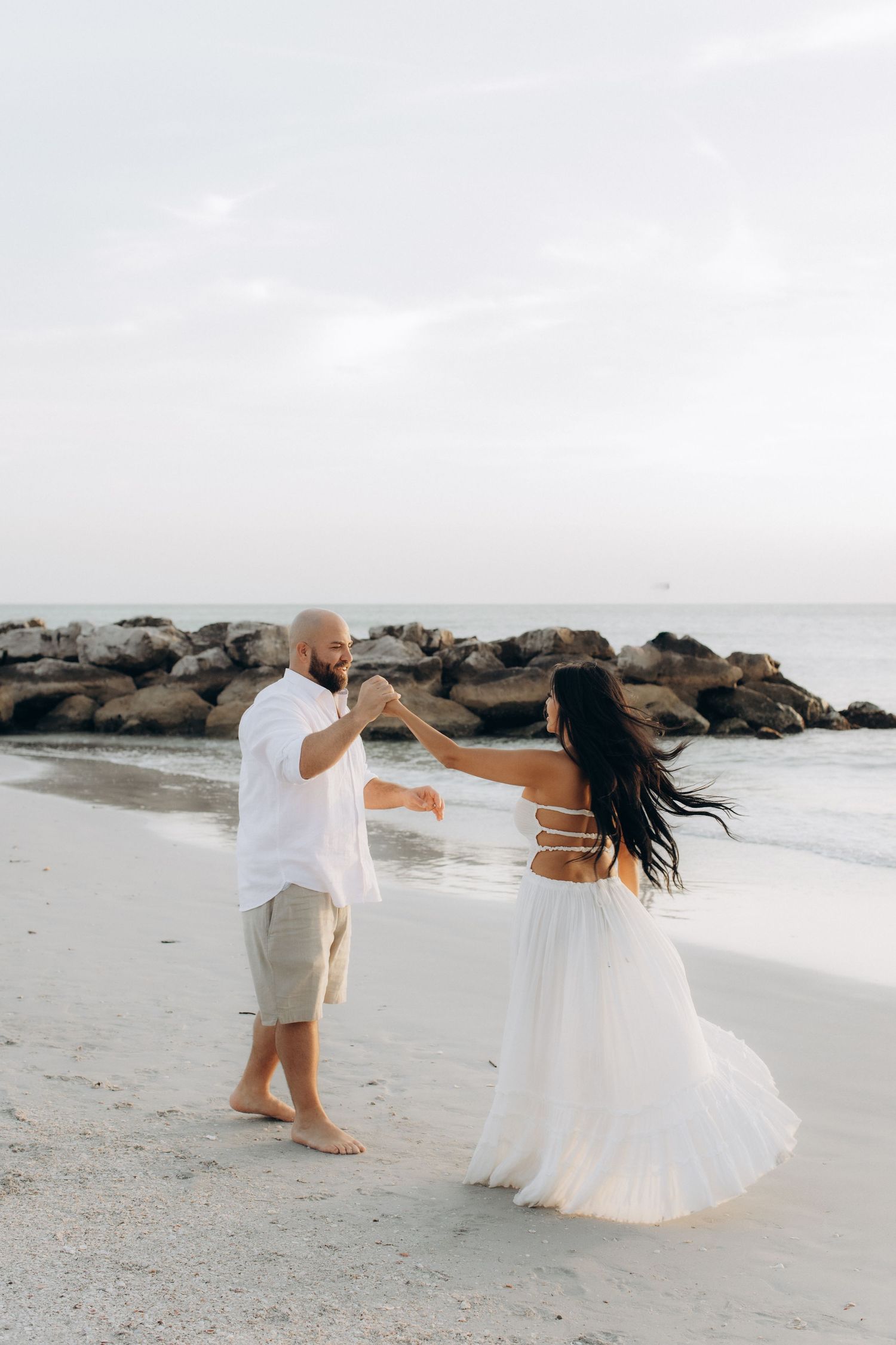 Couple in wedding attire dancing and twirling on a beach with rocky coastline.