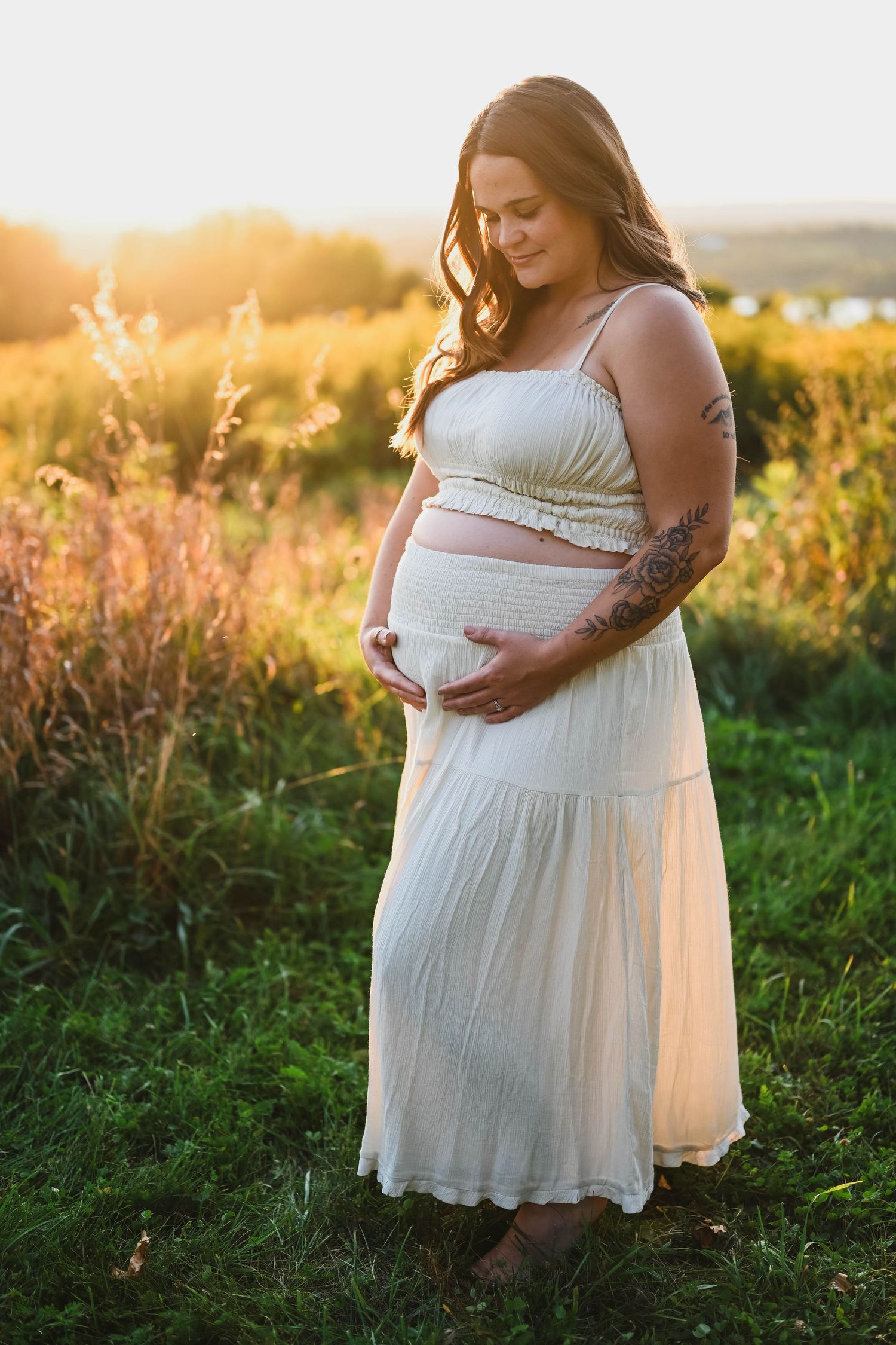 White gown, tattooed mom holding her belly during a maternity session at golden hour with K. Manchuck Photography