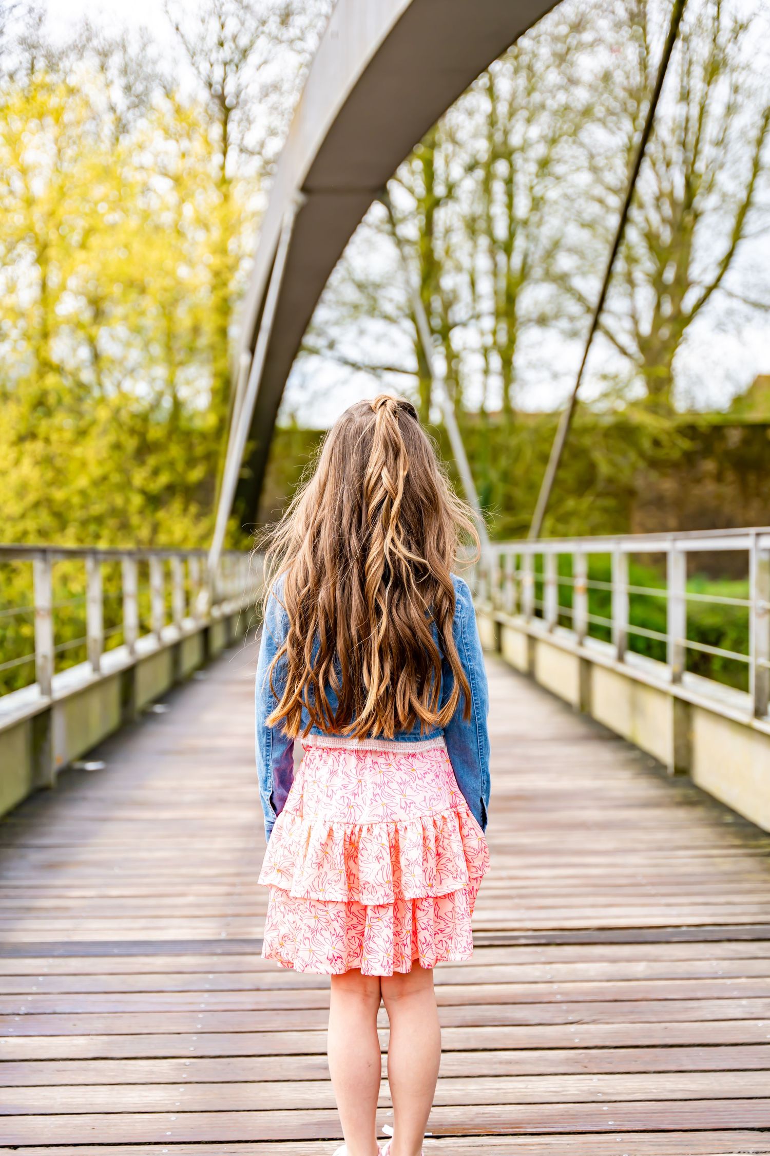 Back view of person in denim jacket and pink skirt standing on wooden bridge.