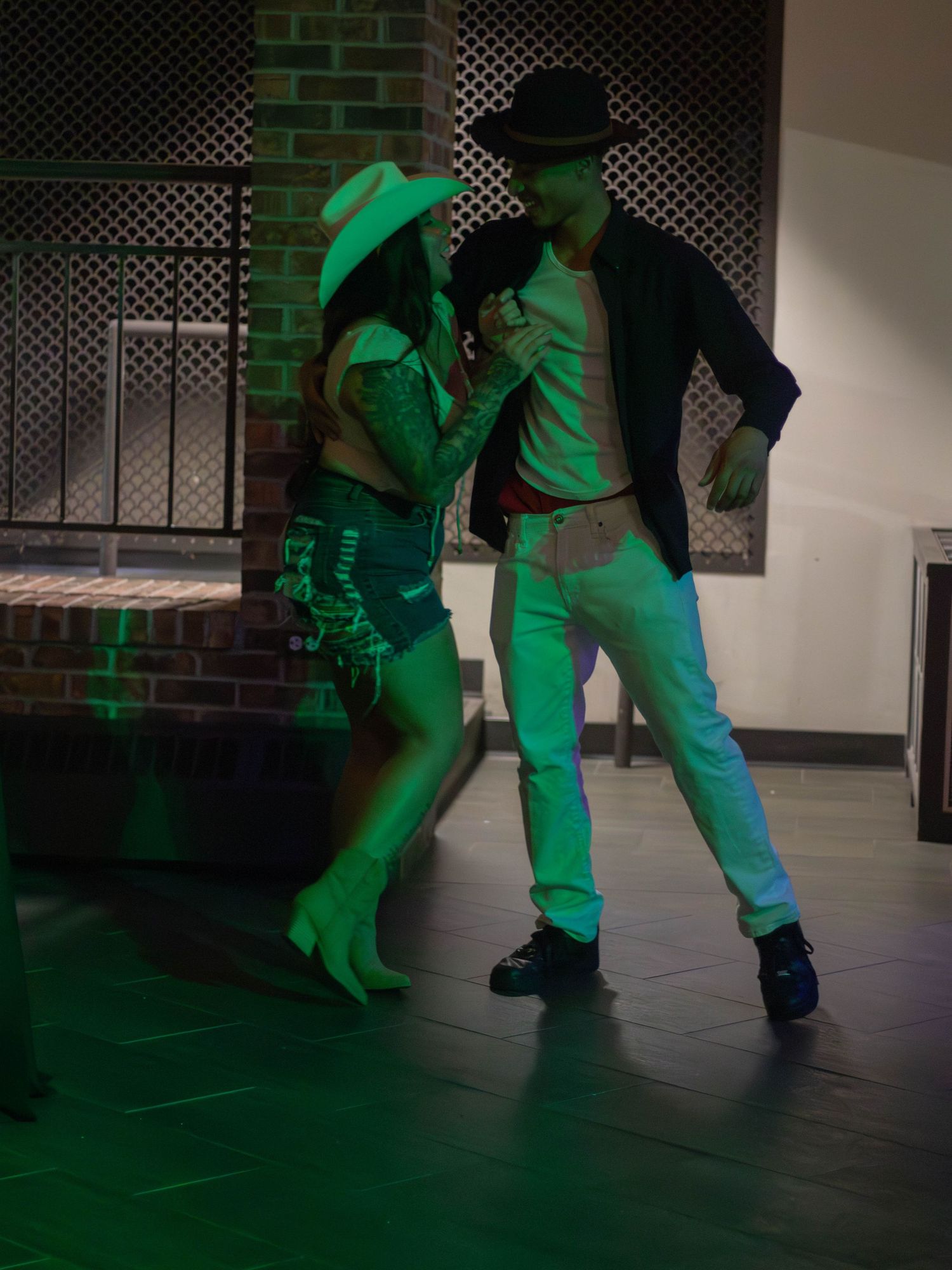 Two dancers performing under green lighting against brick wall background.