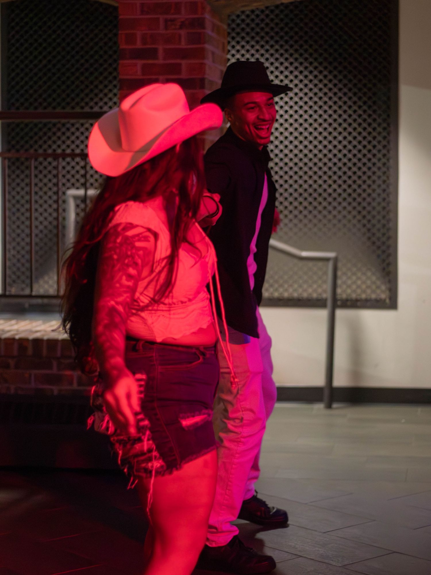 Dancers in western attire illuminated by red lighting perform against brick wall.