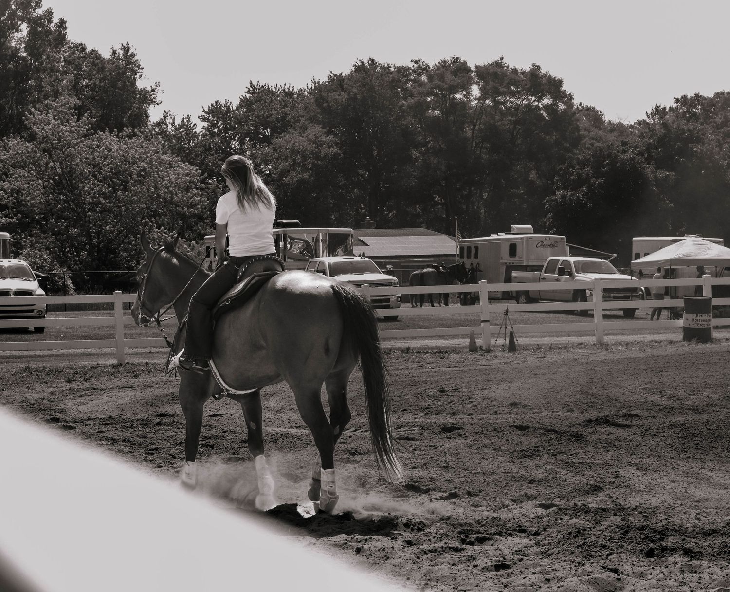 A vintage black and white photo of someone riding a dark horse in an outdoor paddock area.