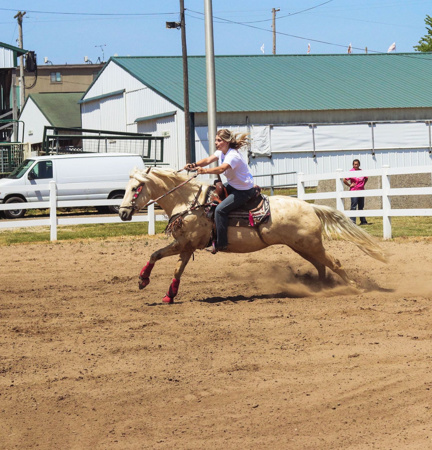 A rider gallops on a palomino horse in a dirt arena with white fencing and buildings in the background.