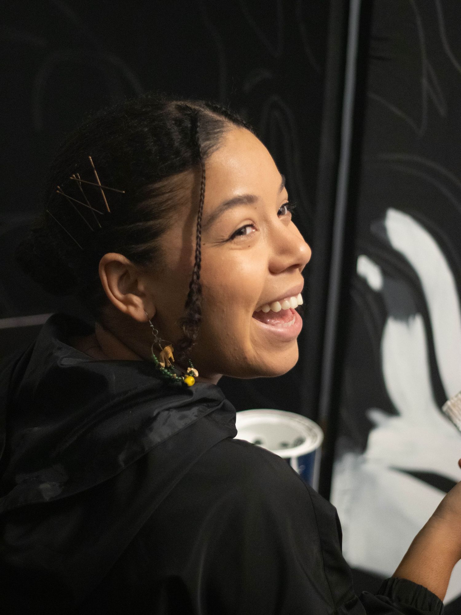 Someone smiling while wearing colorful earrings against a dark background.