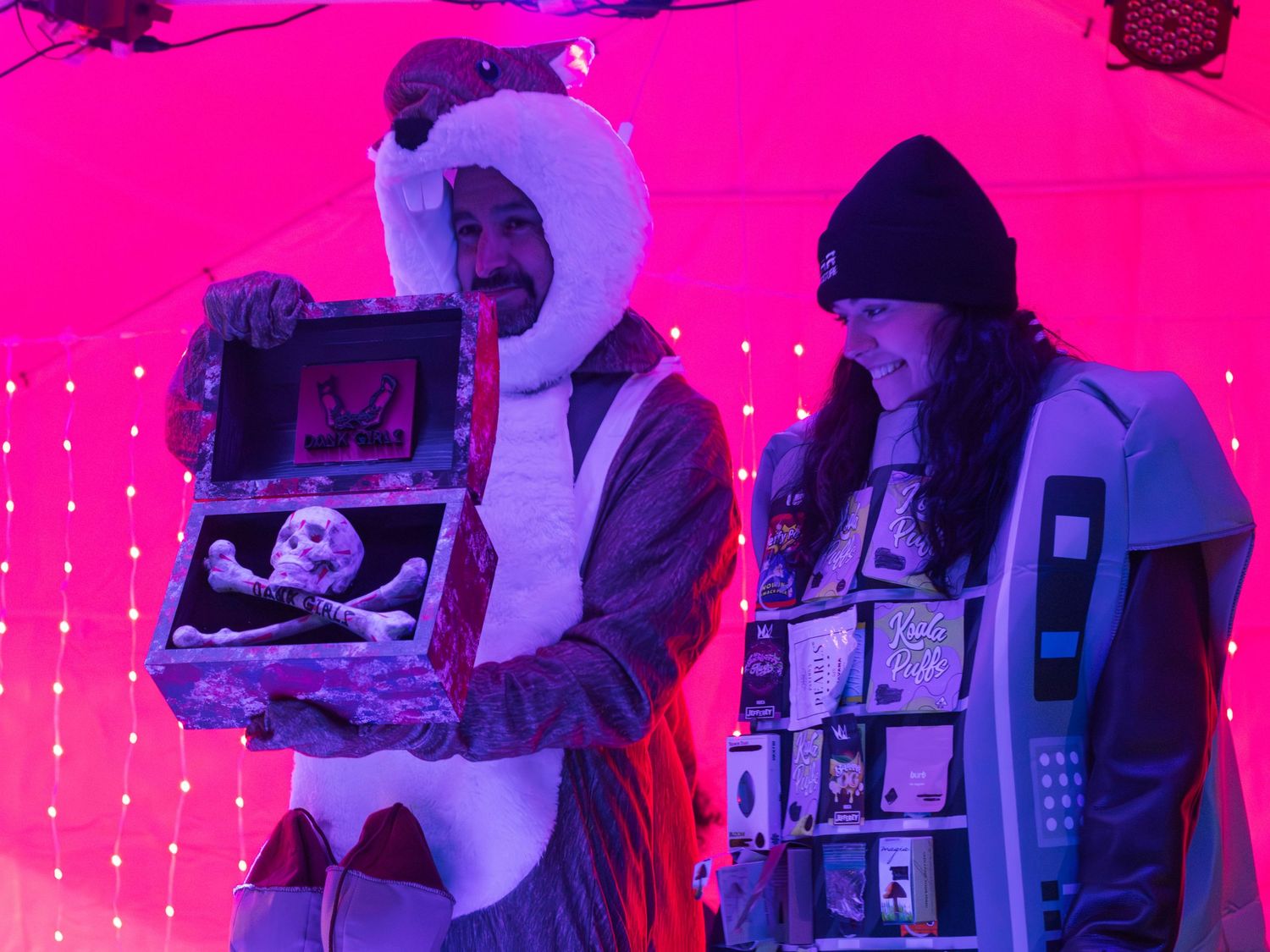 Two people in winter clothing standing under bright pink lighting holding display items.