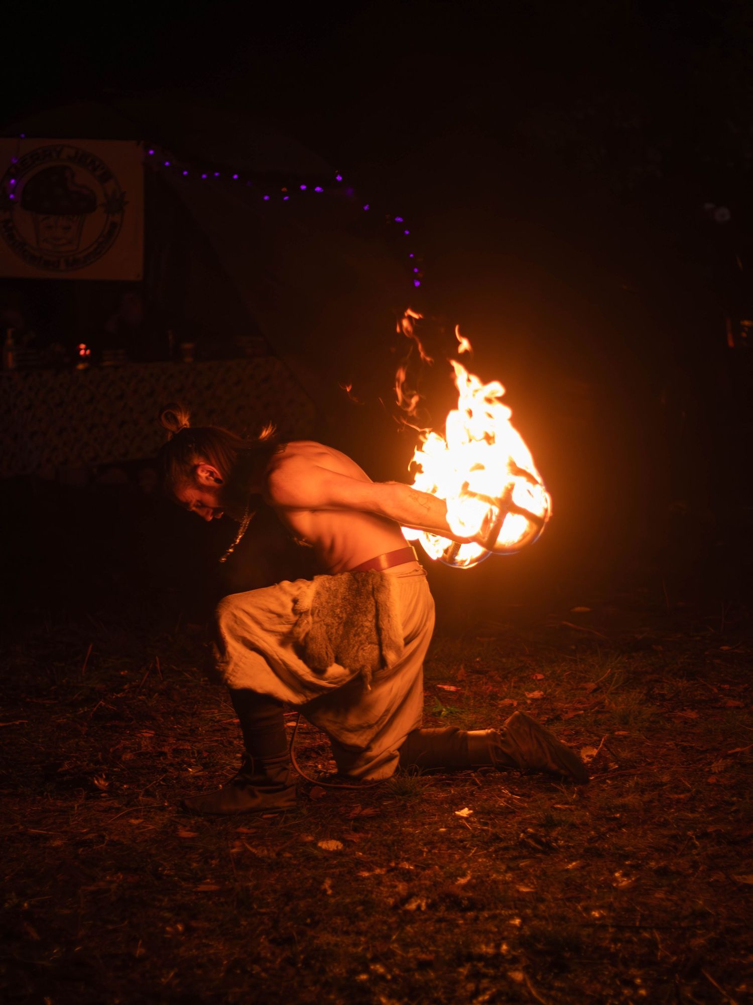 Fire dancer in flowing pants performs low spinning move with flame props at night.