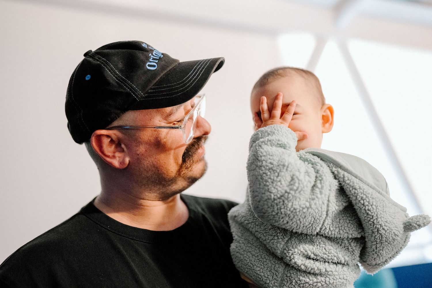 Person wearing a black baseball cap holding a baby wrapped in a grey blanket.