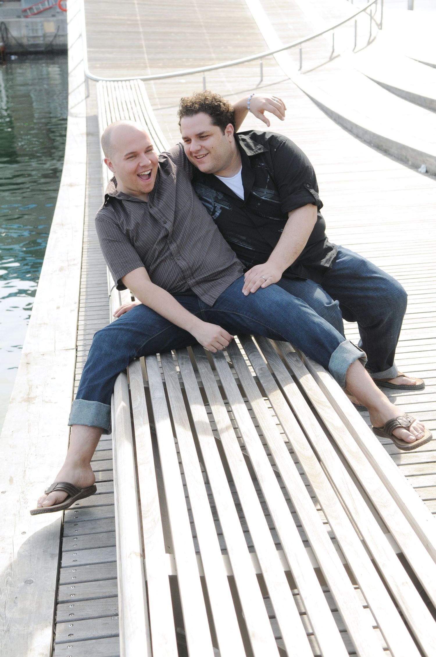 Two people relaxing and laughing together on a wooden bench by the water.