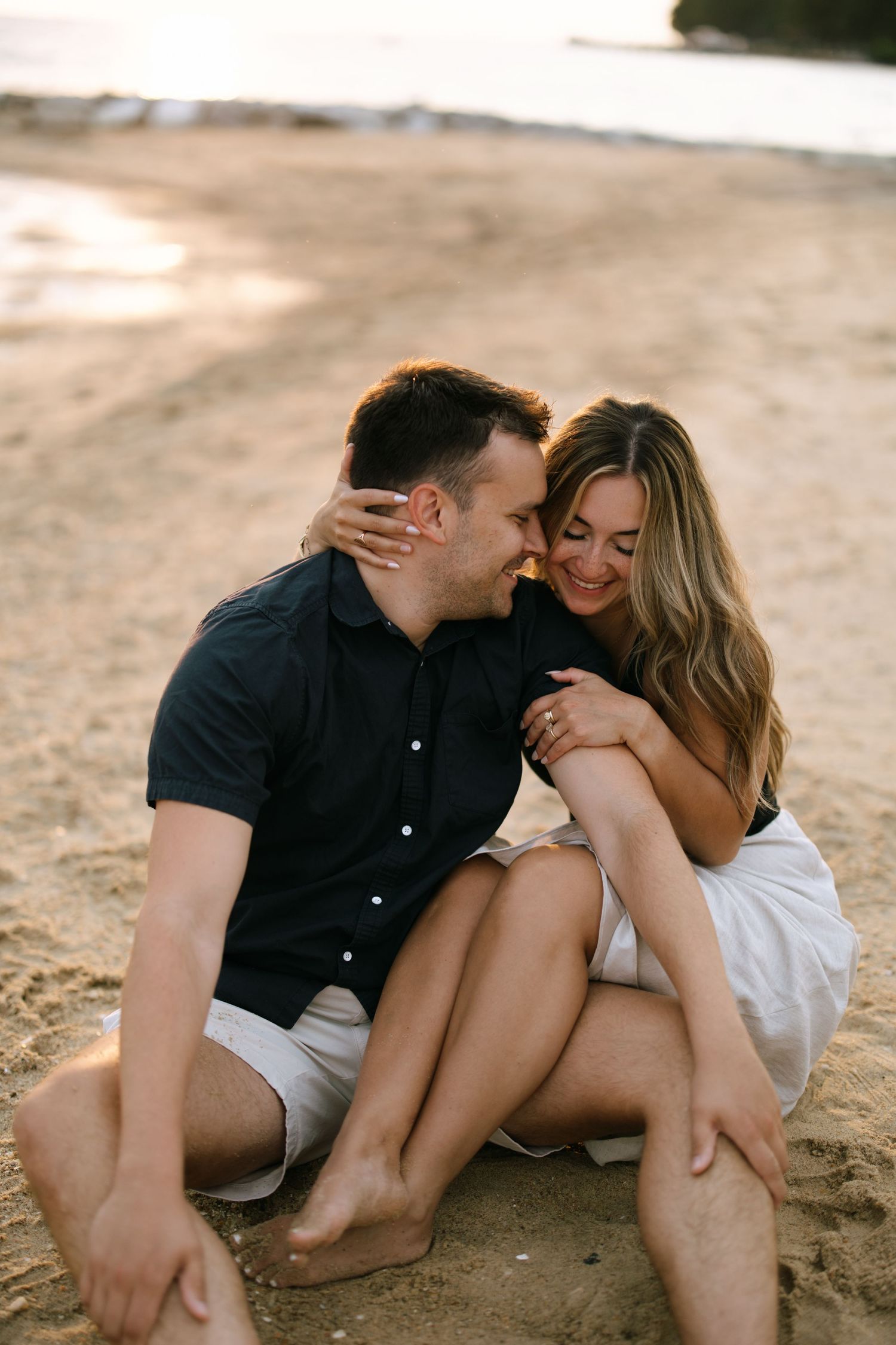 Couple sitting on sandy beach at sunset, smiling and embracing, woman in white skirt.