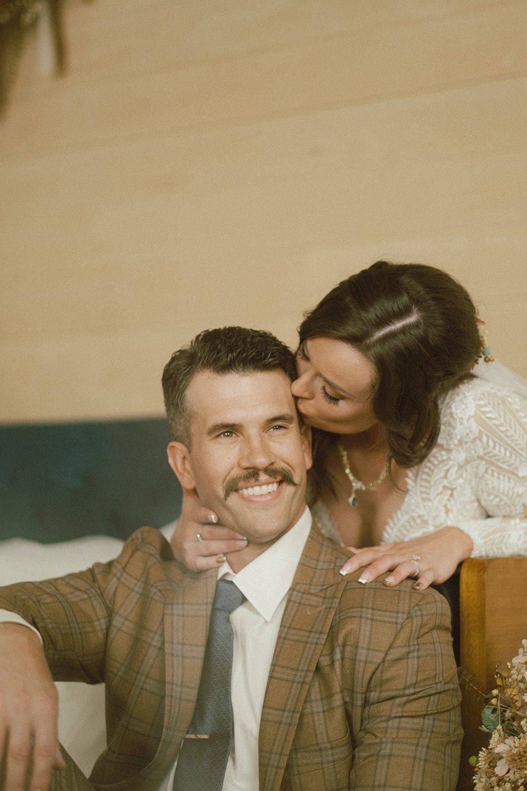 Smiling groom in plaid suit with bride kissing his cheek, indoor wedding portrait.
