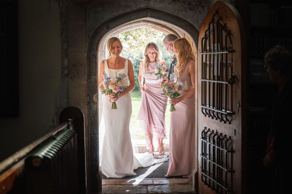 A wedding party in white and pink dresses stand in an arched stone entryway holding bouquets.