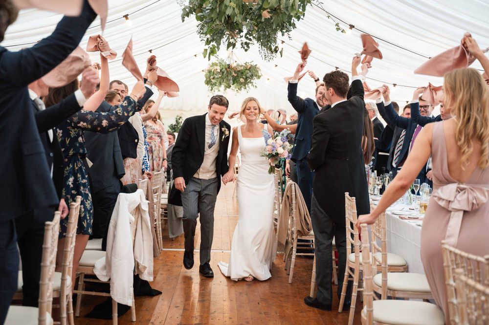 Celebratory scene of wedding guests cheering in a decorated marquee as couple walks down the aisle.