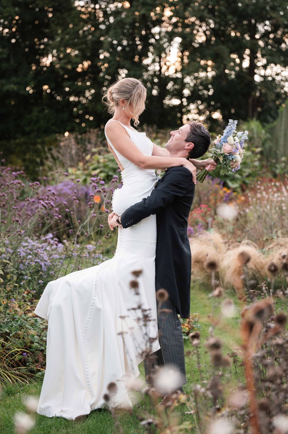 A joyful wedding moment captured as couple poses playfully in a garden filled with late summer flowers.