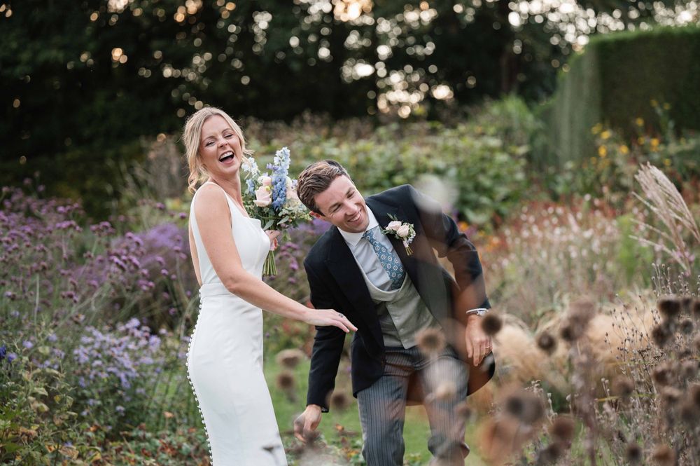 A joyful wedding couple laughs together in a garden filled with purple wildflowers at sunset.