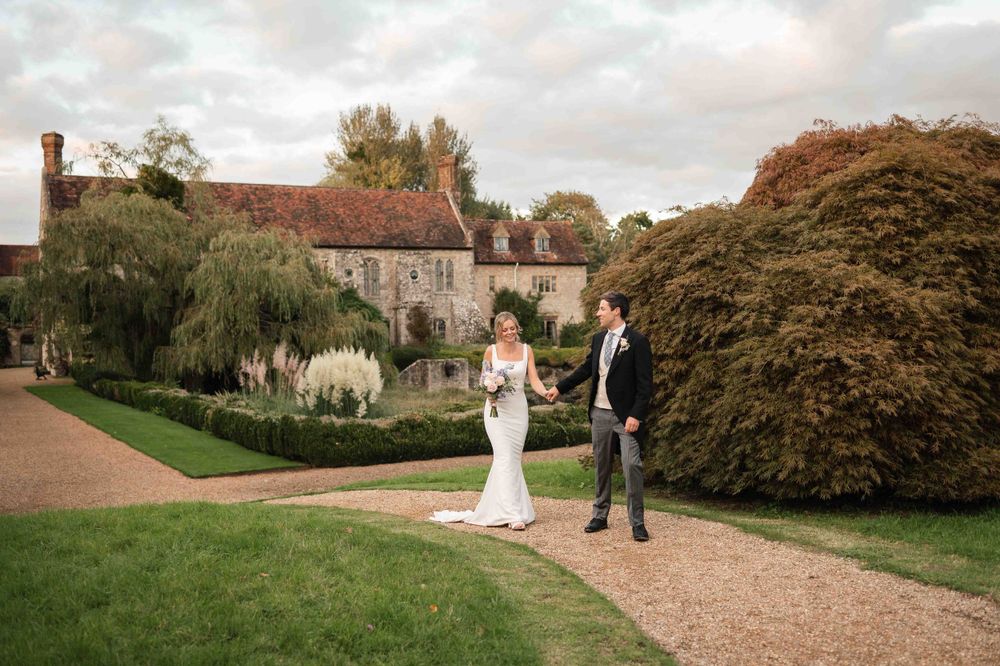 Two people in formal attire walk along a gravel path at an elegant country estate with manicured gardens and stone buildings.