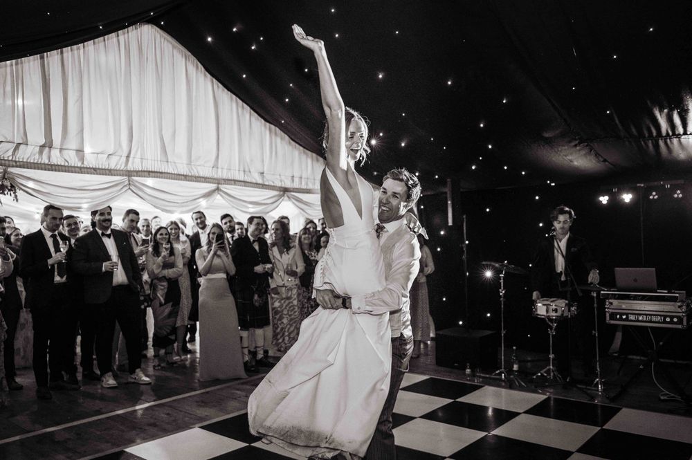 A couple performs a graceful first dance on a checkerboard dance floor while guests look on at an elegant wedding reception.