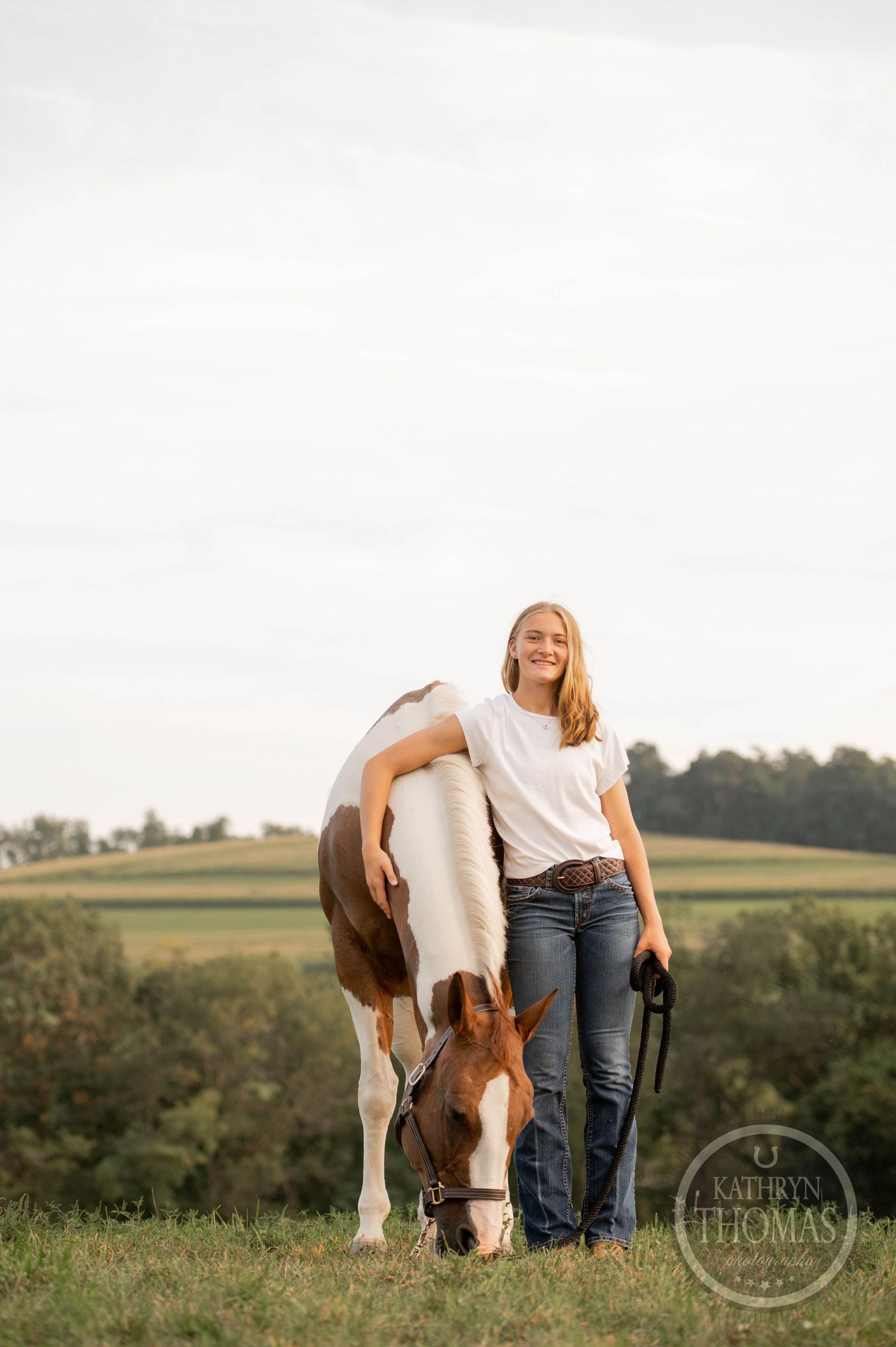 Person stands with grazing paint horse against scenic rural landscape at sunset.
