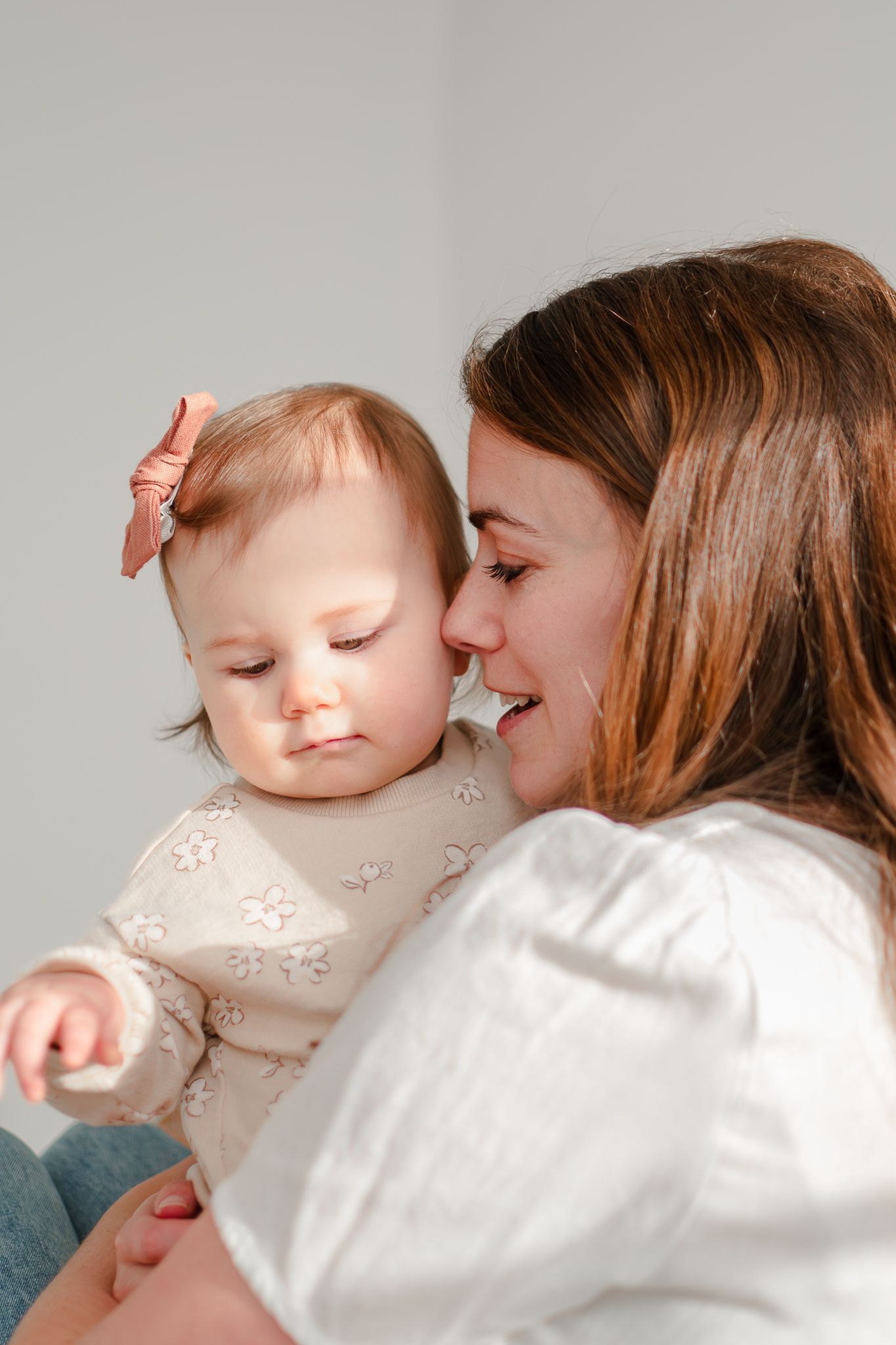 A sweet moment of affection and laughter between a mother and baby in white against a neutral background.