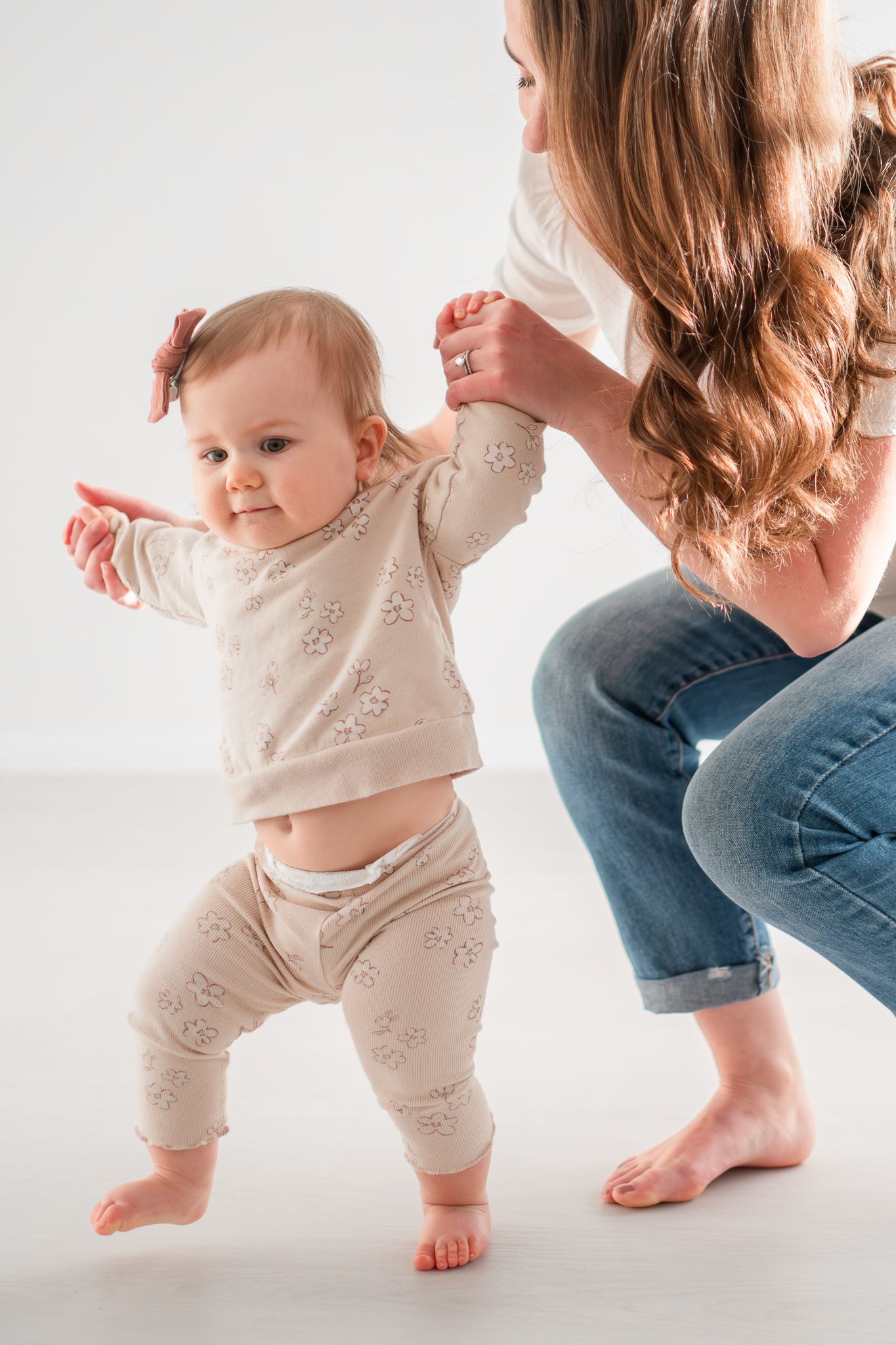 Adorable baby in beige outfit takes first steps with support from parent on bright white background.