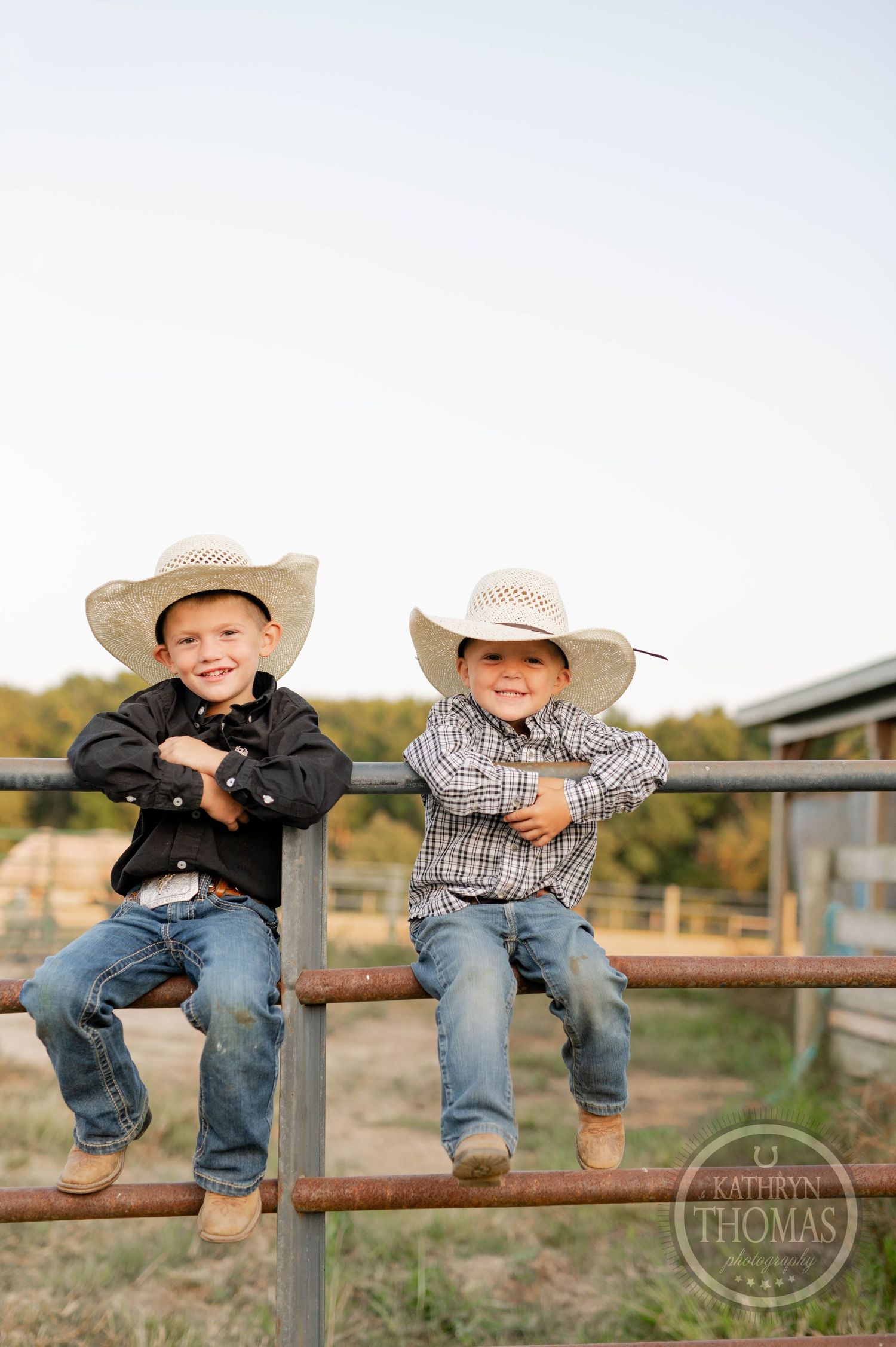 Two young cowboys in straw hats sit together on metal fence rails at a farm.
