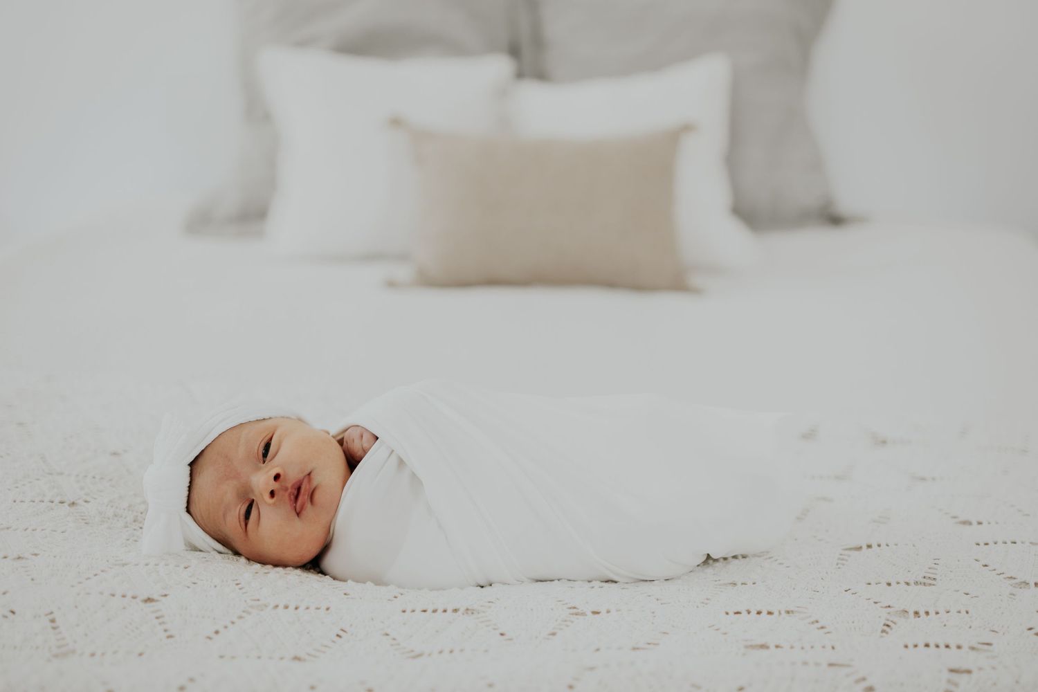 Tiny newborn baby swaddled in white blankets on a white bed with soft beige pillows in background.