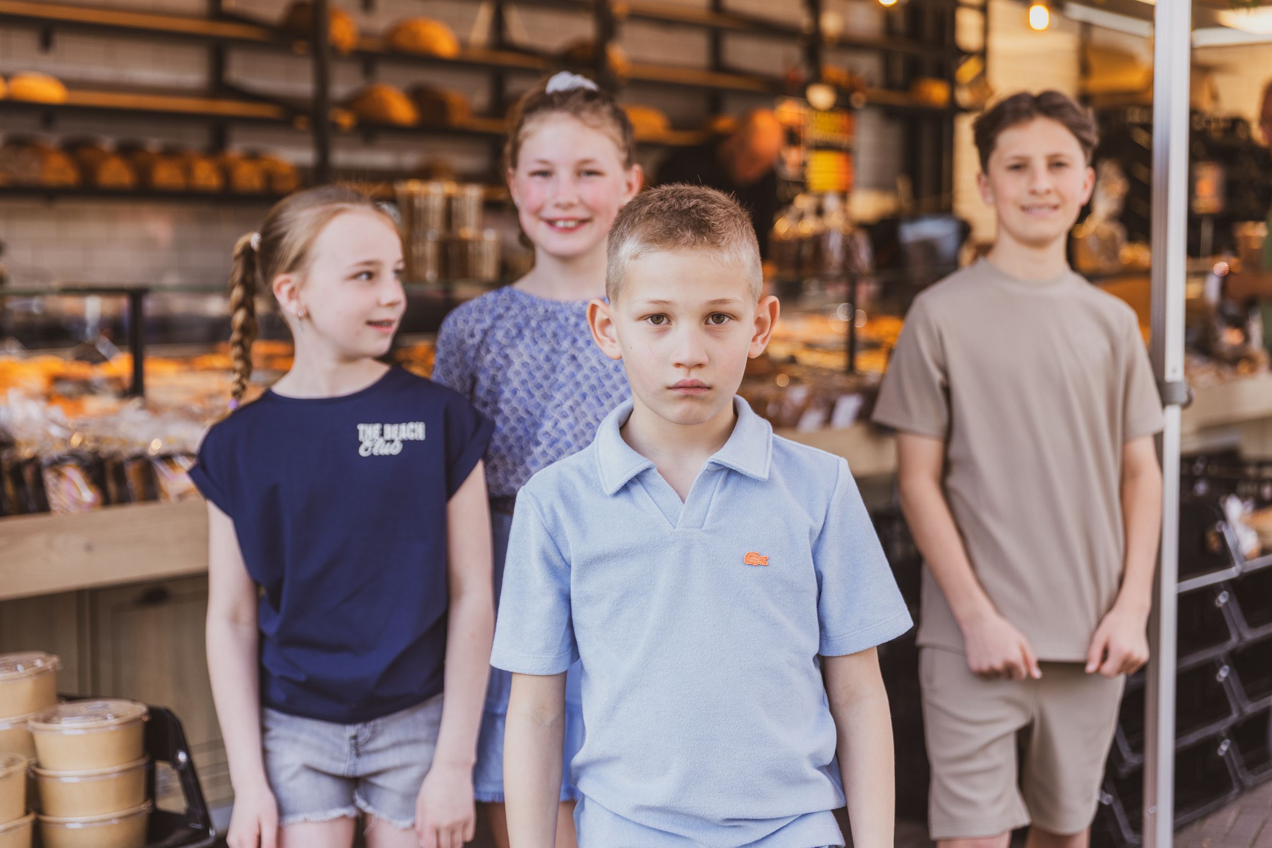 Four children pose together in a bakery shop, with shelves of bread visible in the warm background.