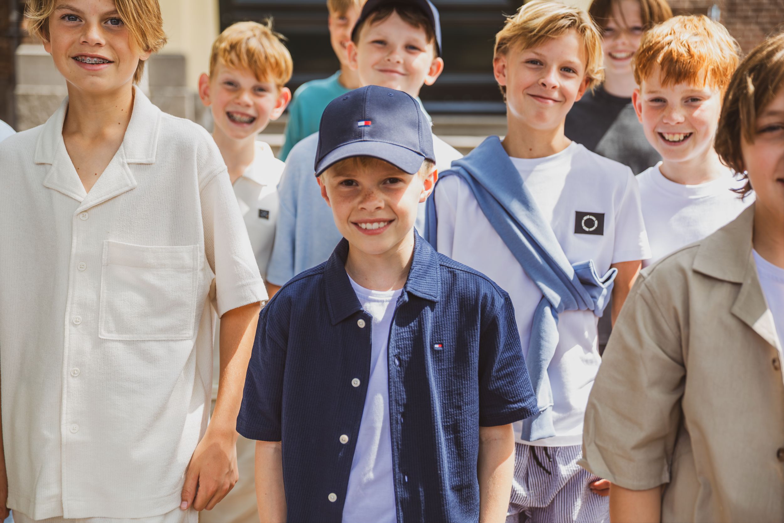 A group of smiling boys wearing casual summer clothing, with one boy in a navy shirt and blue cap standing in front.