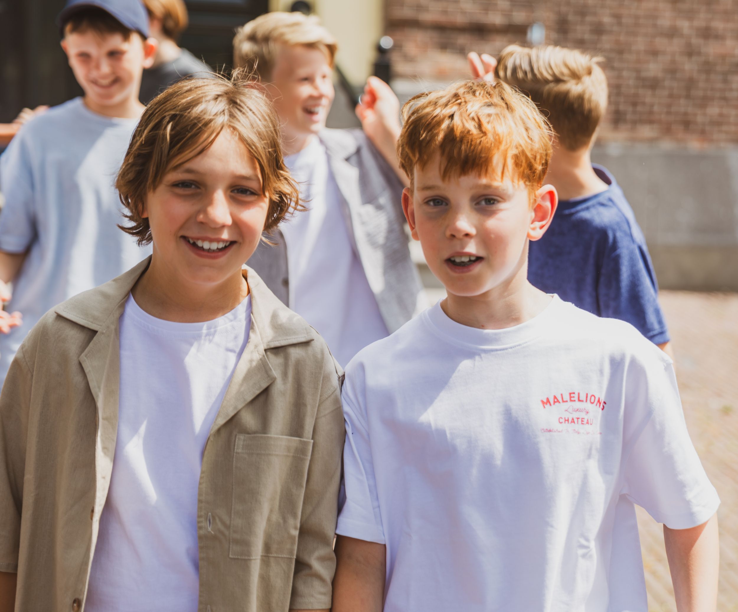 Two smiling boys in white t-shirts stand together outdoors, with other children visible in the background.