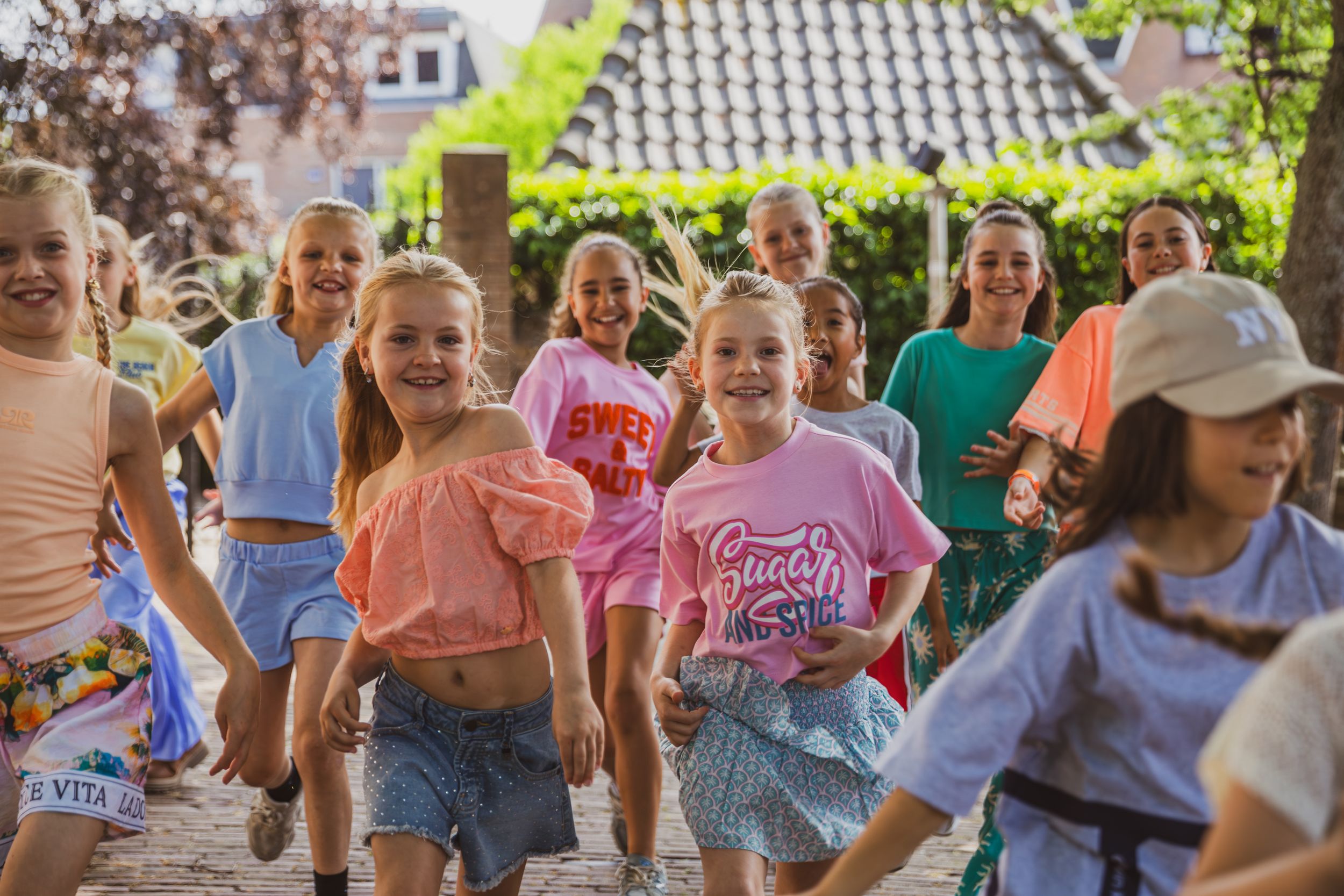 Group of happy girls running and laughing outdoors in a sunny courtyard, wearing colorful summer clothes.