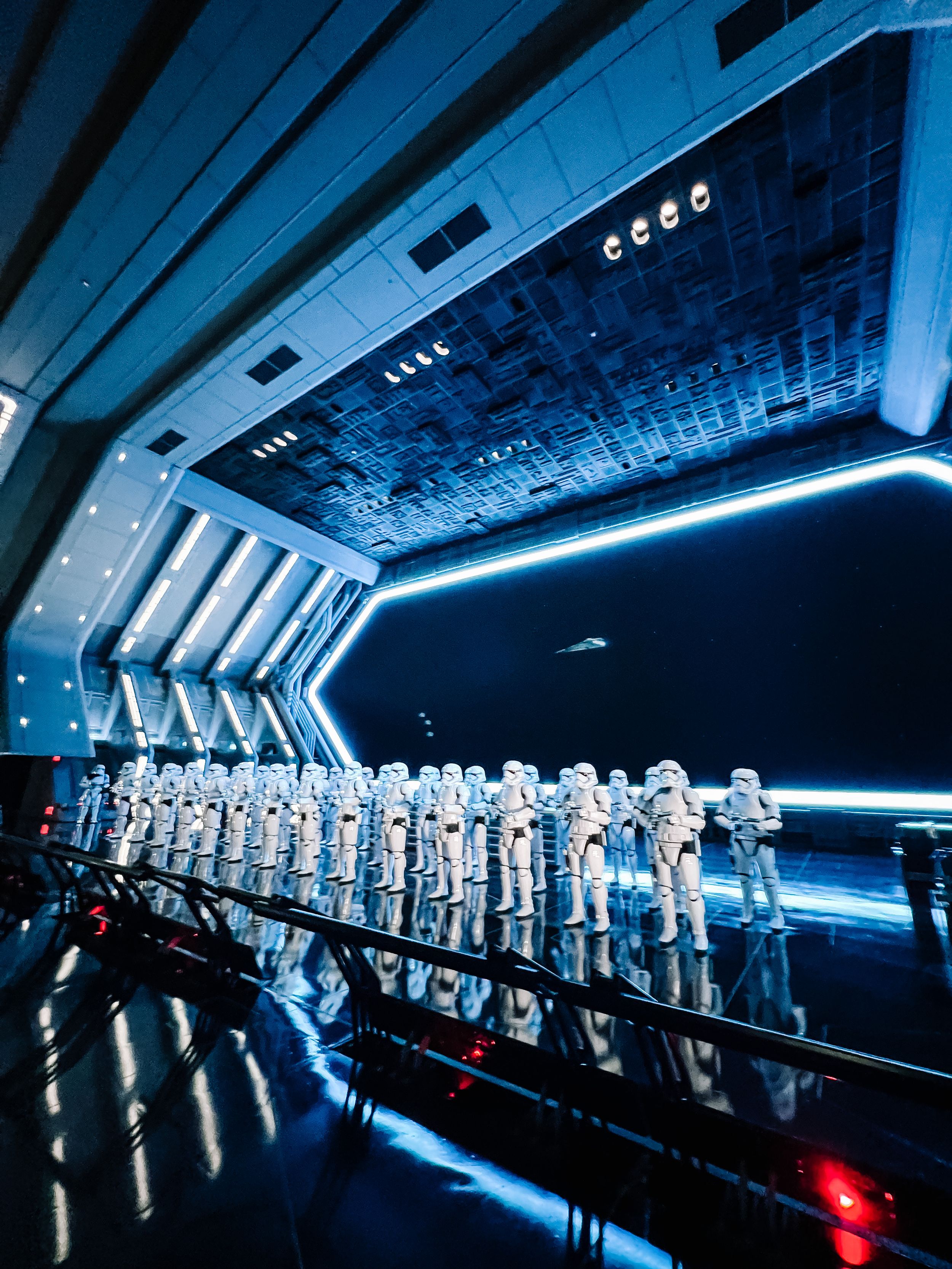 A futuristic exhibit space with rows of white armored costumes lined up in a sleek blue-lit corridor with reflective floors.