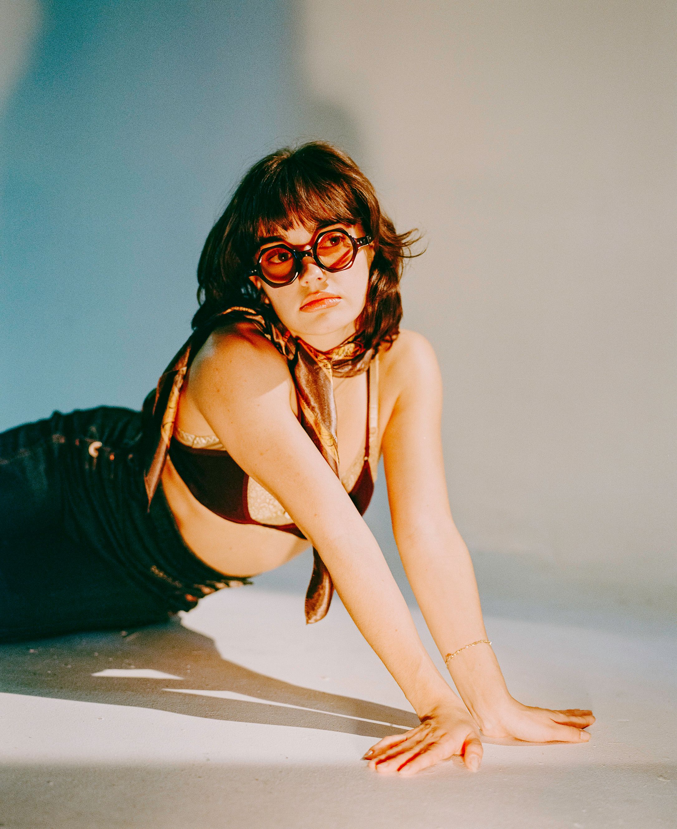 Fashion portrait of a model in sunglasses and dark swimwear posing on a light floor.