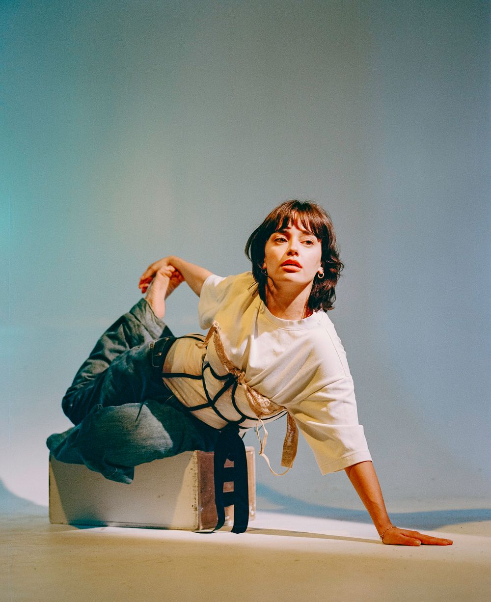 Modern dance pose with figure crouching in white and beige outfit against turquoise background.