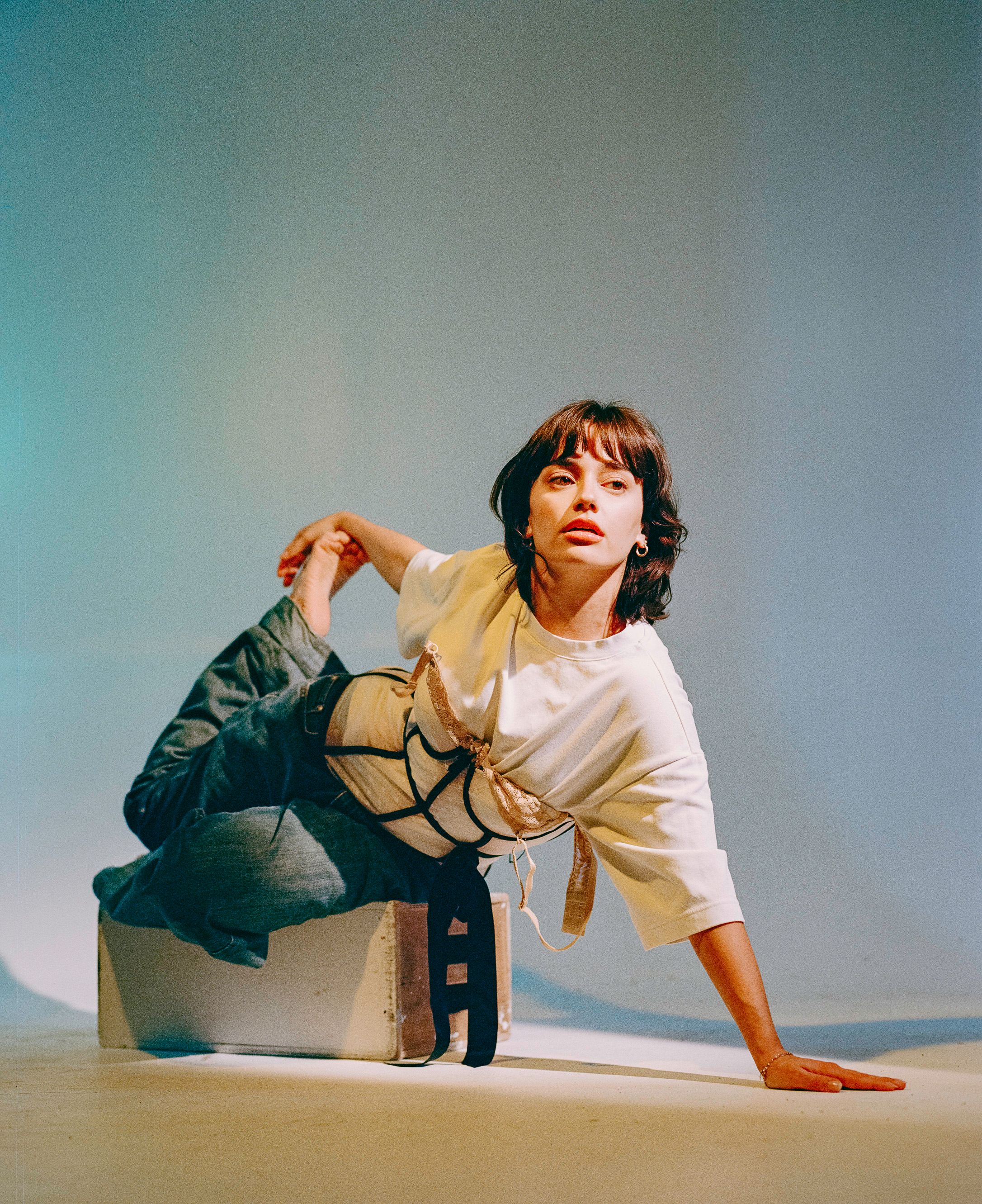 Modern dance pose with figure crouching in white and beige outfit against turquoise background.