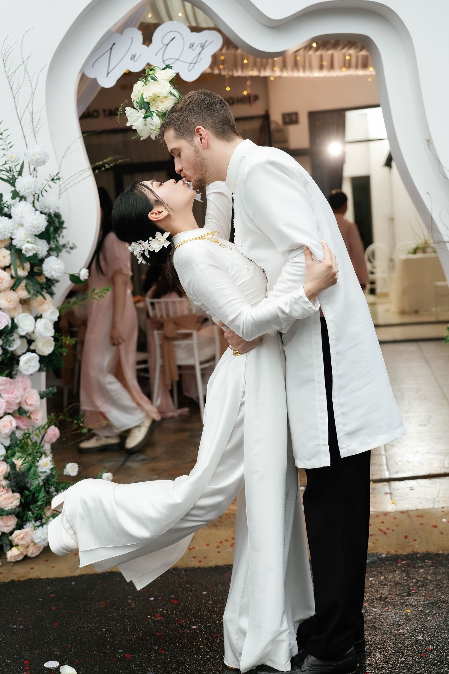 A romantic wedding moment under a floral arch as the couple shares a kiss while the bride's dress flows elegantly.