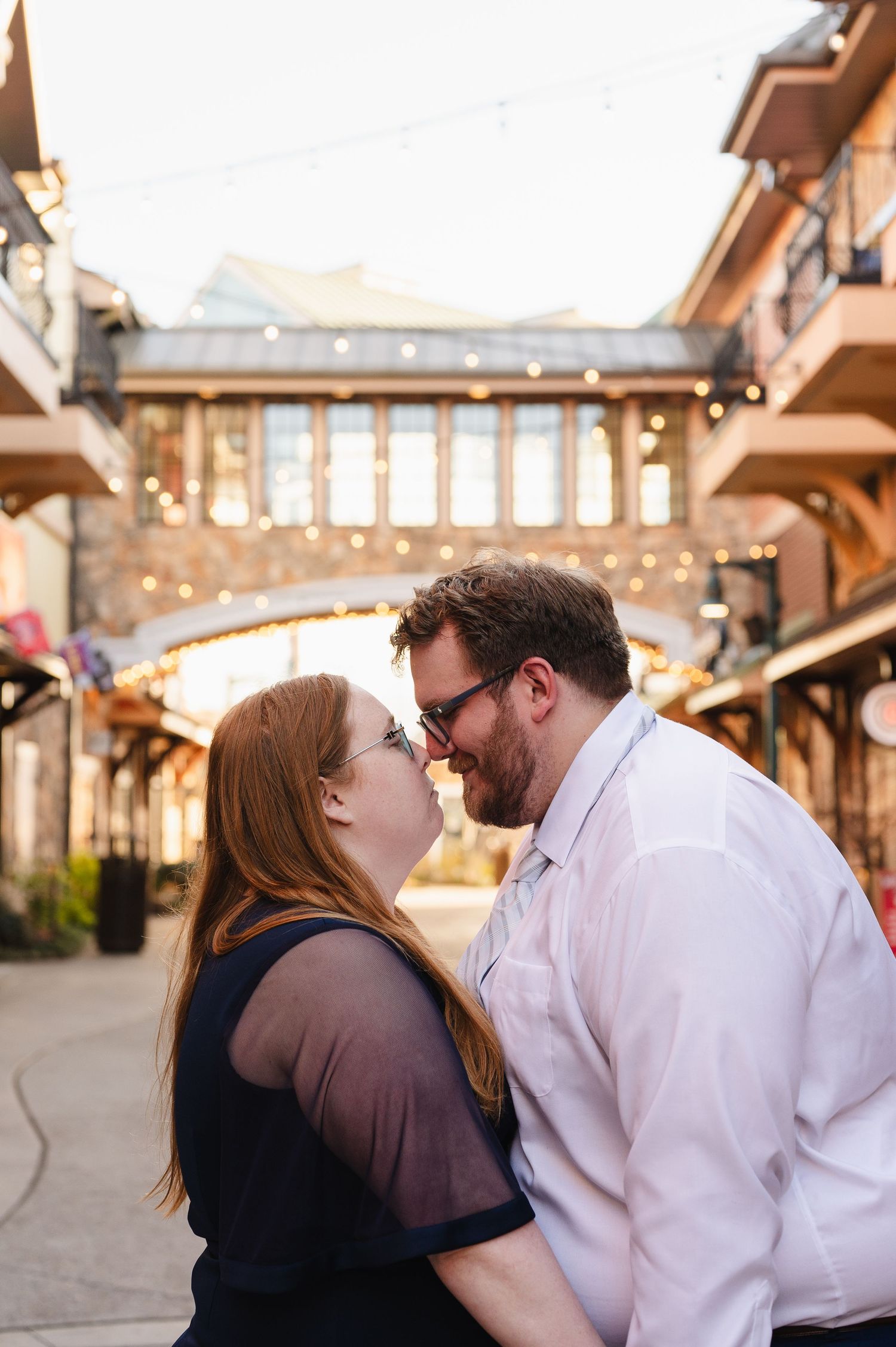A couple shares a romantic moment under a covered bridge walkway decorated with string lights at sunset.