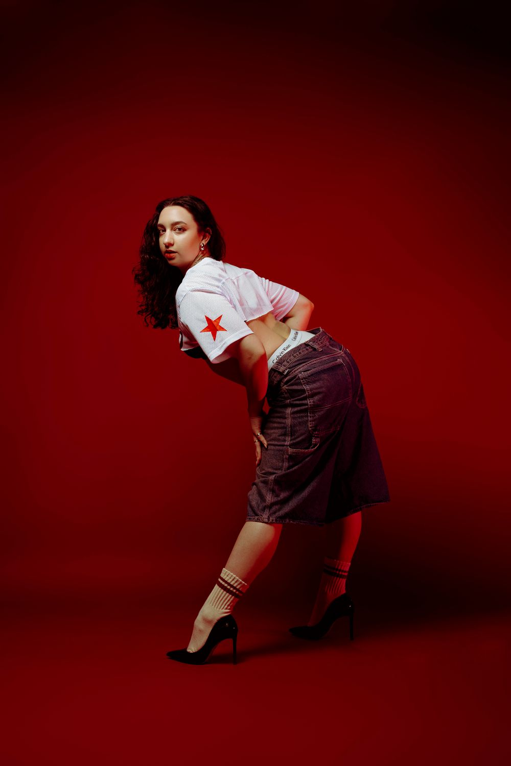 Fashion model strikes dynamic pose in Tokyo jersey and cargo pants against red backdrop.