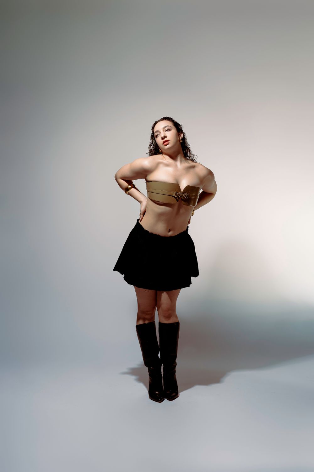 Fashion model in black miniskirt and knee-high boots poses confidently against light studio background.