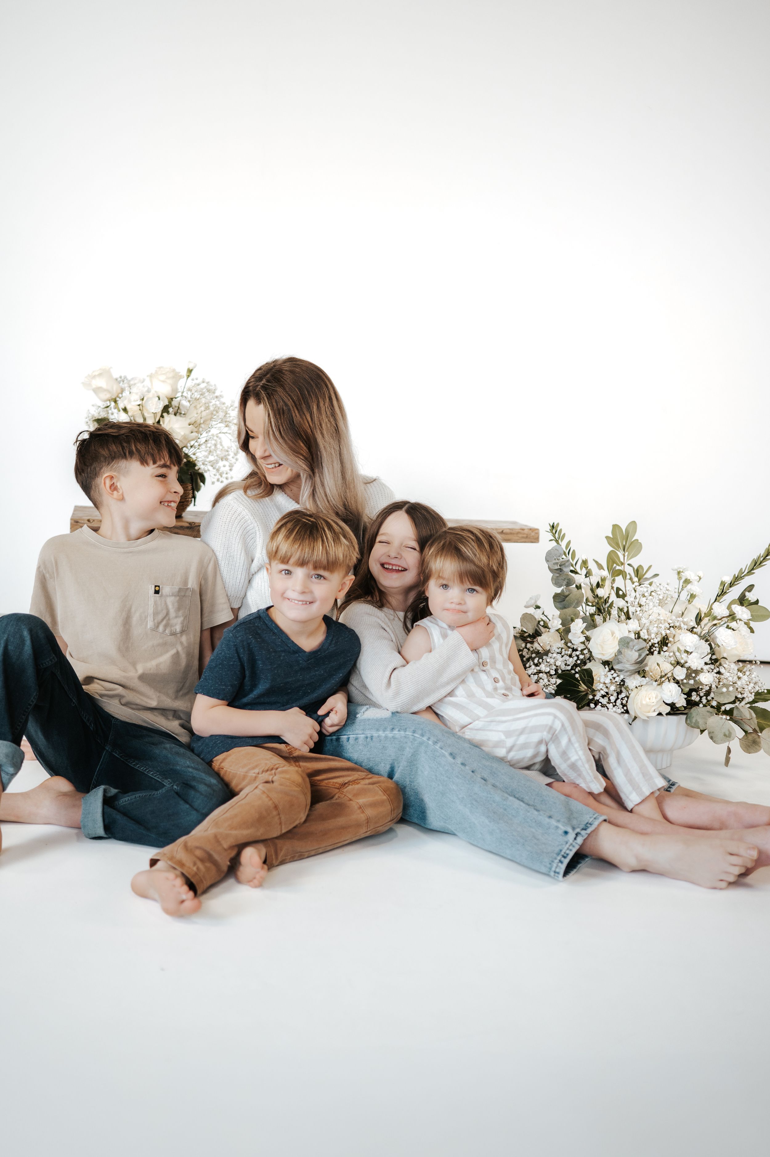 A loving family group sits together in casual clothes against a white backdrop with delicate floral arrangements.