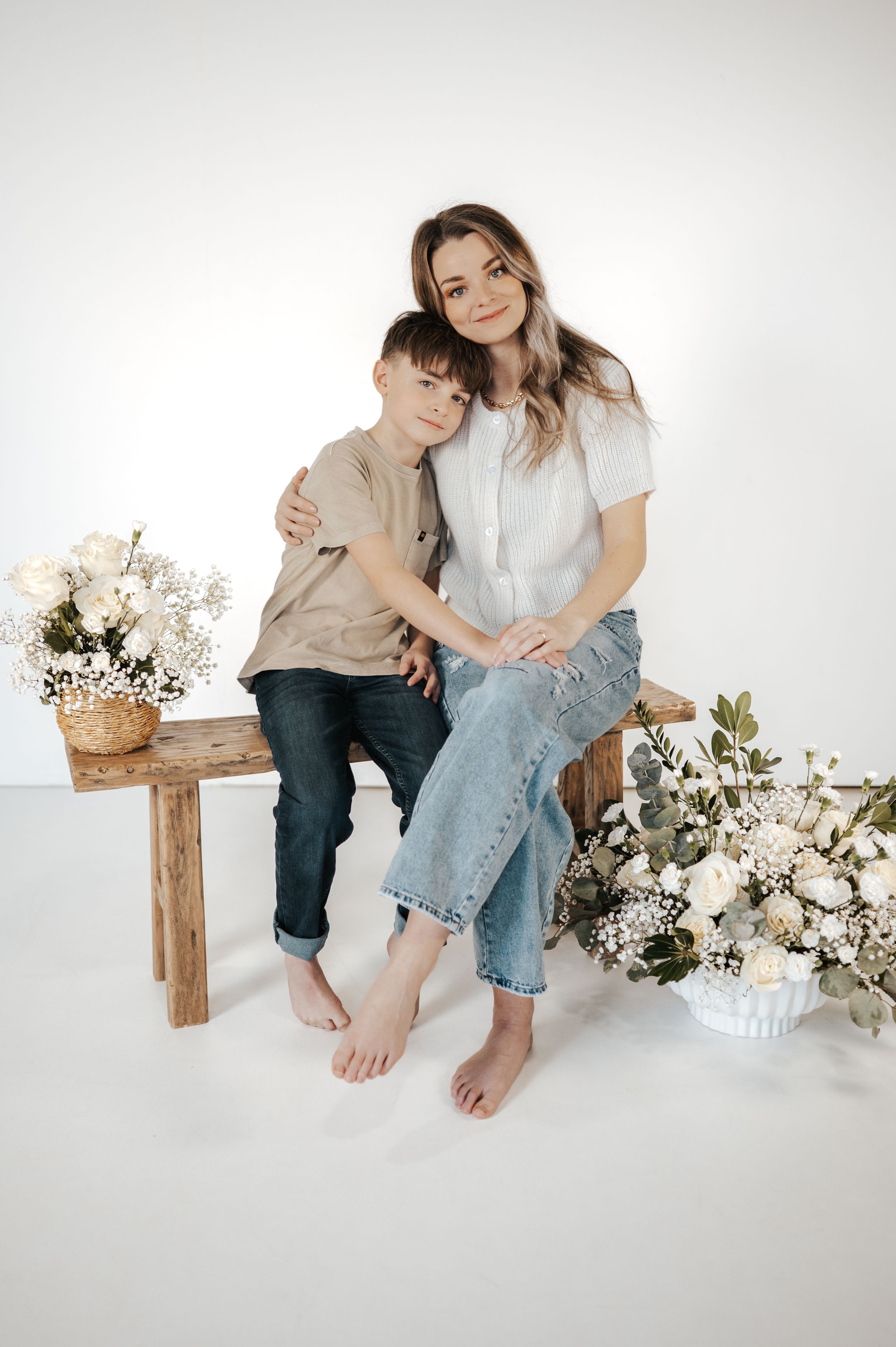 A sweet portrait of two people sitting on a wooden bench surrounded by white floral arrangements.