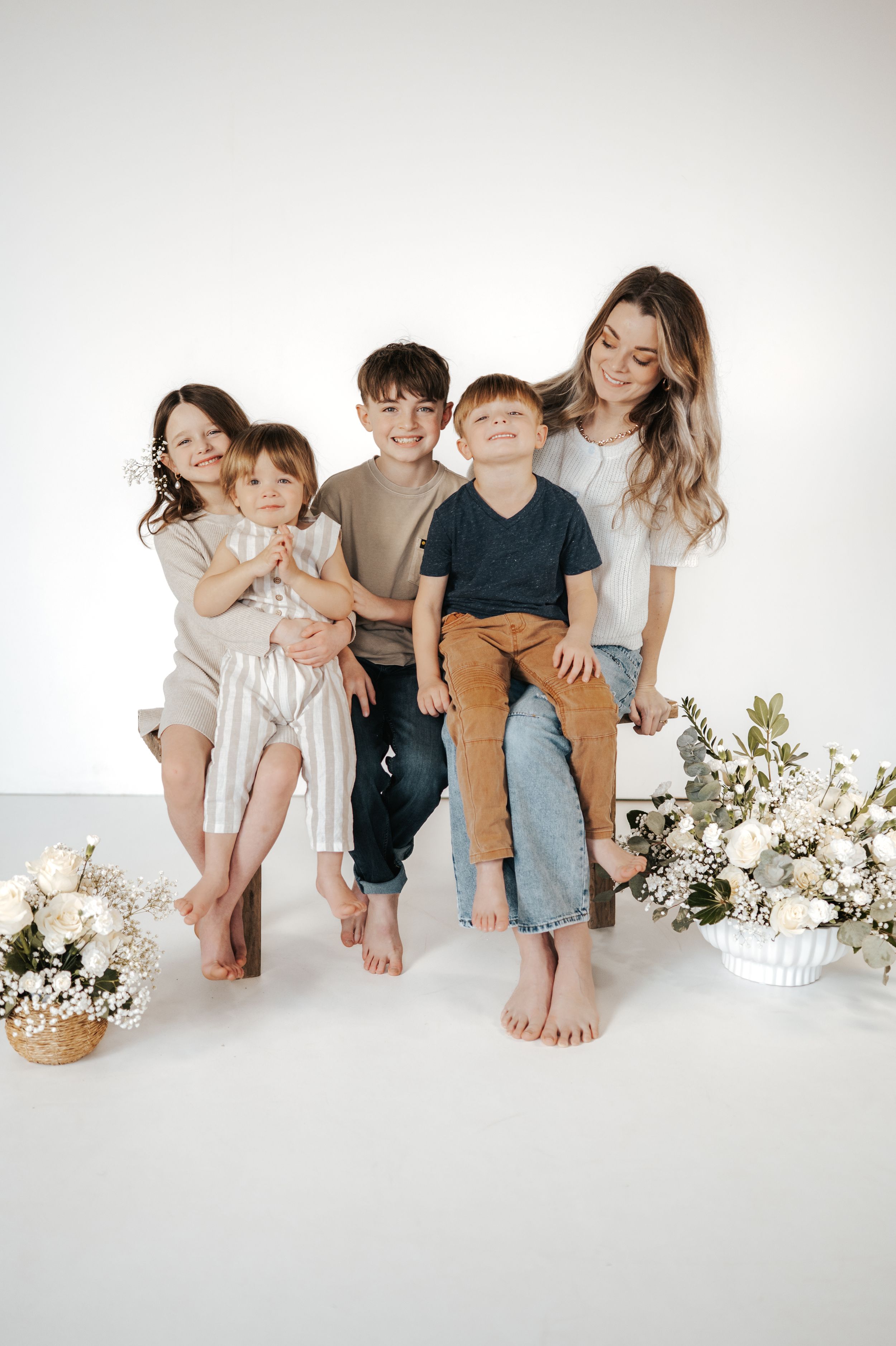 Family portrait with siblings in casual attire sitting together on white backdrop with floral decorations.