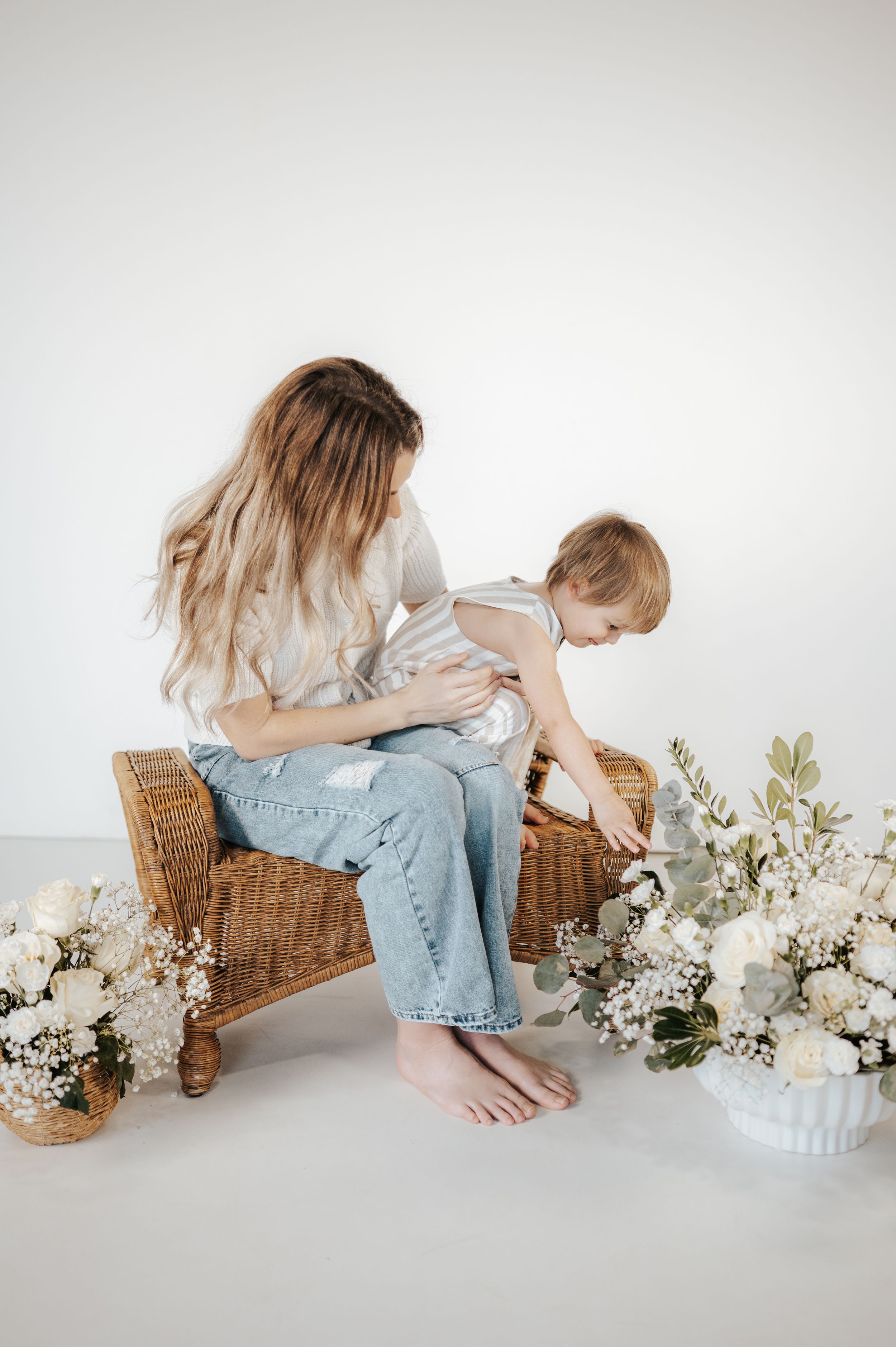 Tender parenting moment captured in minimalist setting with wicker chair and white floral arrangements.