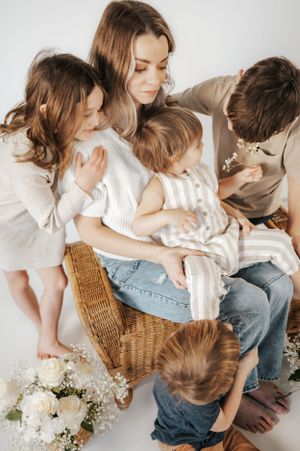 Family in matching cream sweaters gather on wicker chair with white floral bouquet beside them.
