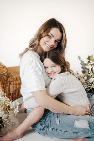 Parent and child in matching white sweaters share a tender hug while sitting against a light background with dried flowers.